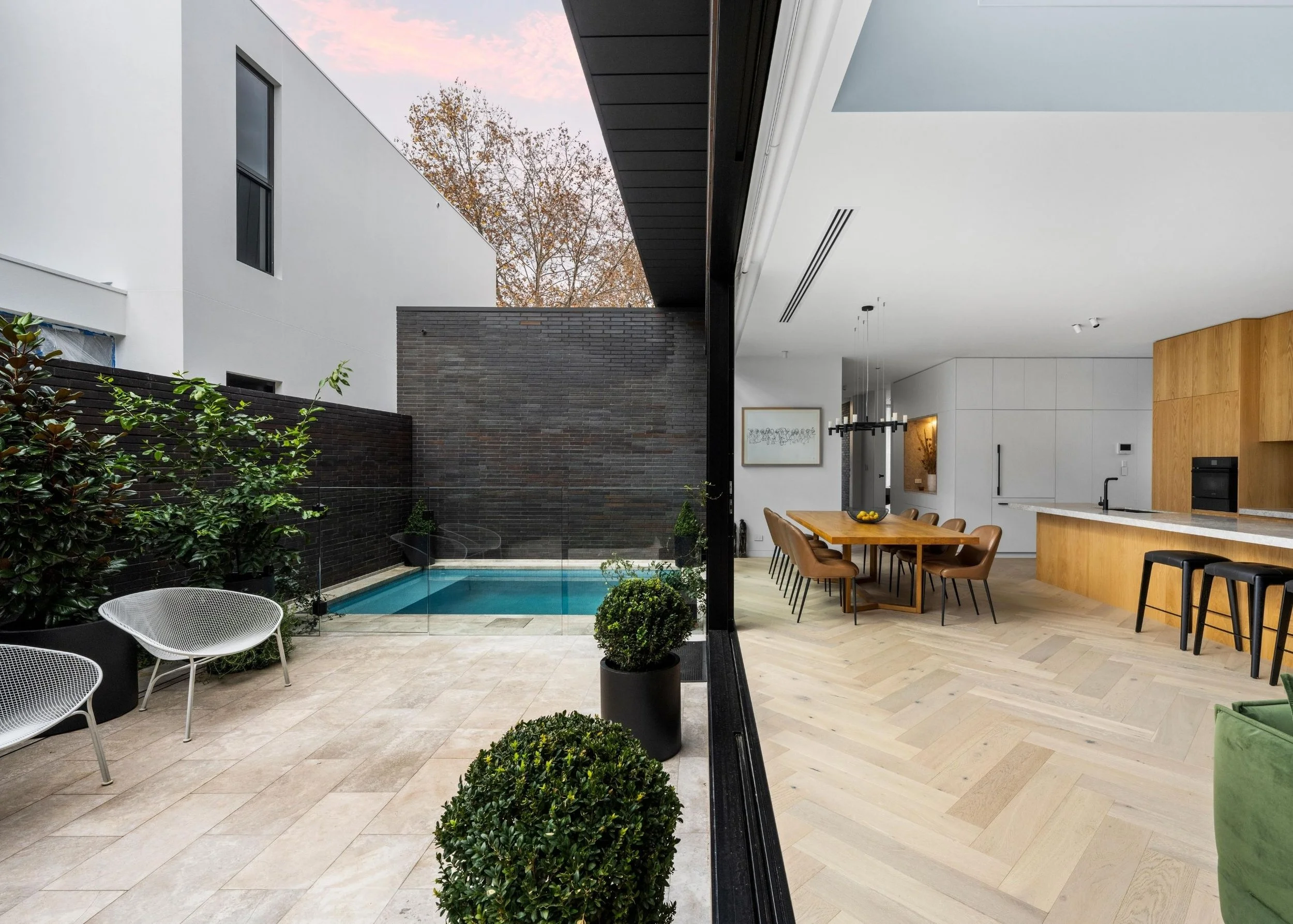 Modern house interior with open kitchen and dining area, large windows, and a view of an outdoor pool and patio with planters and chairs.
