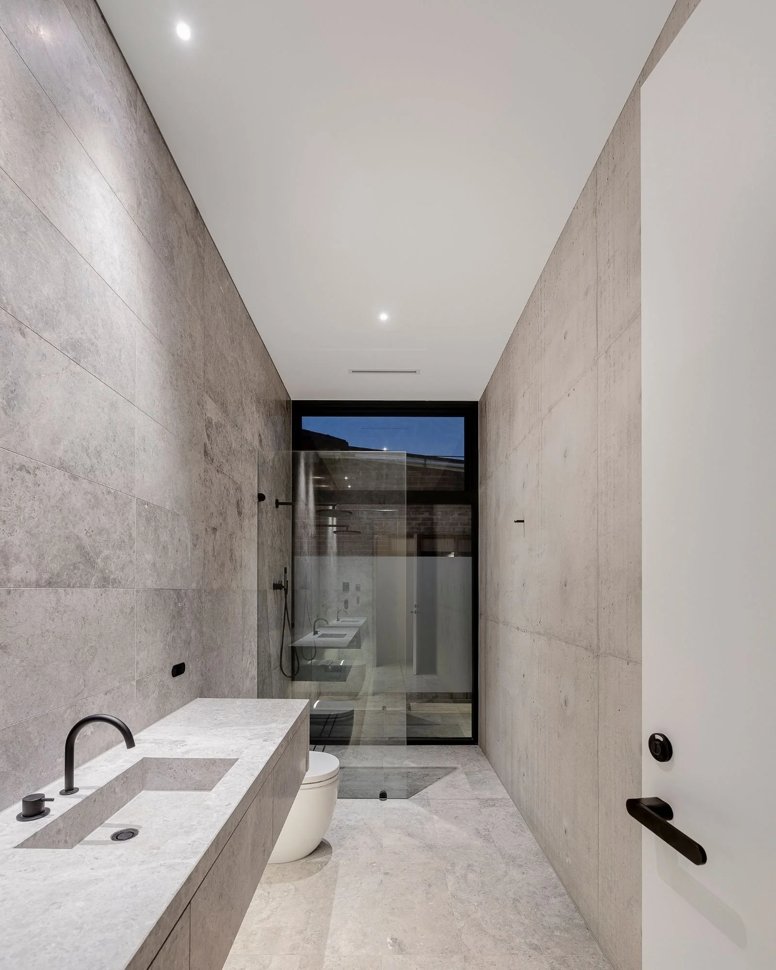 Modern bathroom with beige tiles, a white countertop with a black faucet, a toilet, a walk-in shower with a glass door, and a window showing an outdoor view.