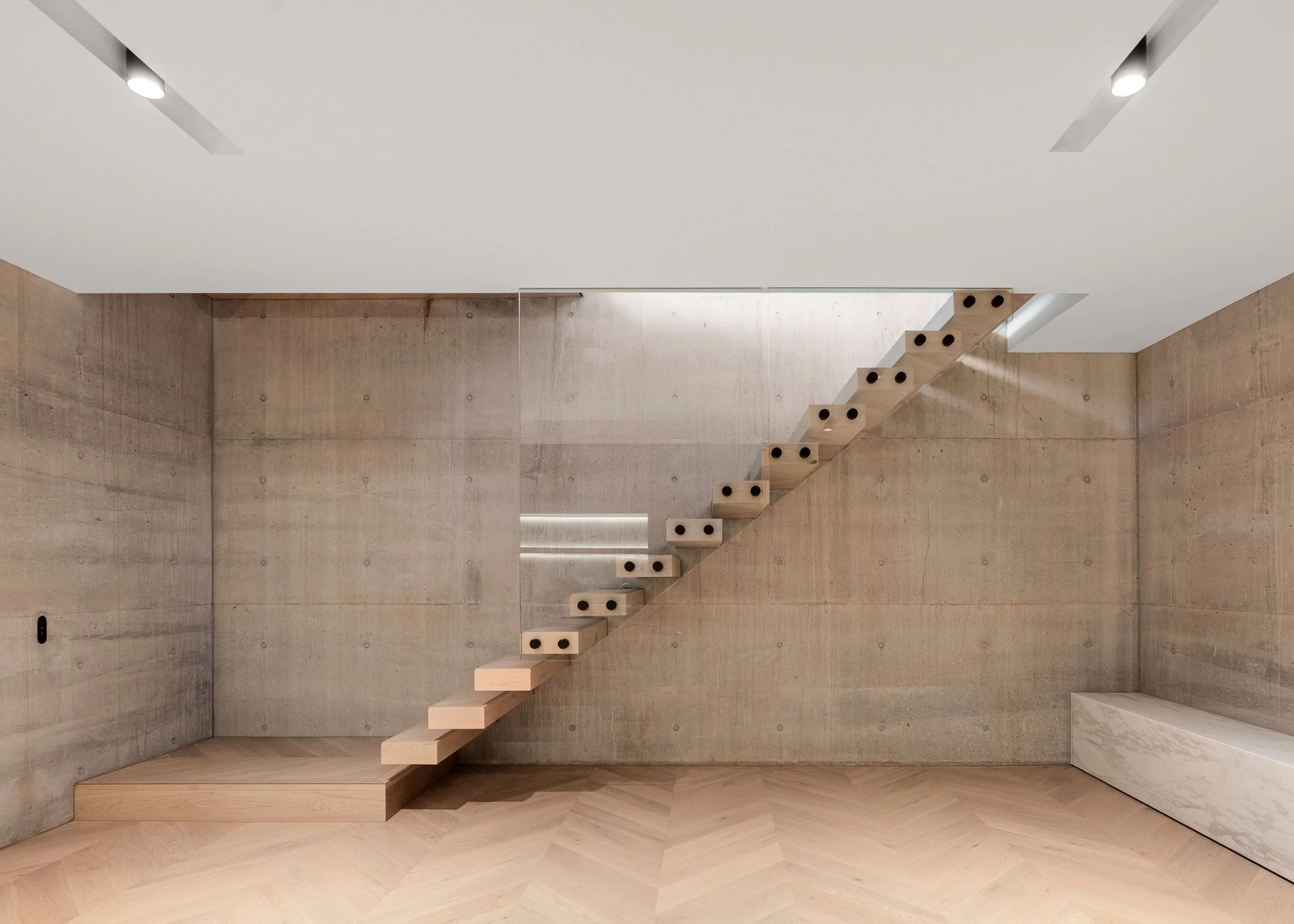 Interior of a modern building featuring a floating staircase with wooden steps and black circular supports, minimalist concrete walls, and a white ceiling with recessed lighting.