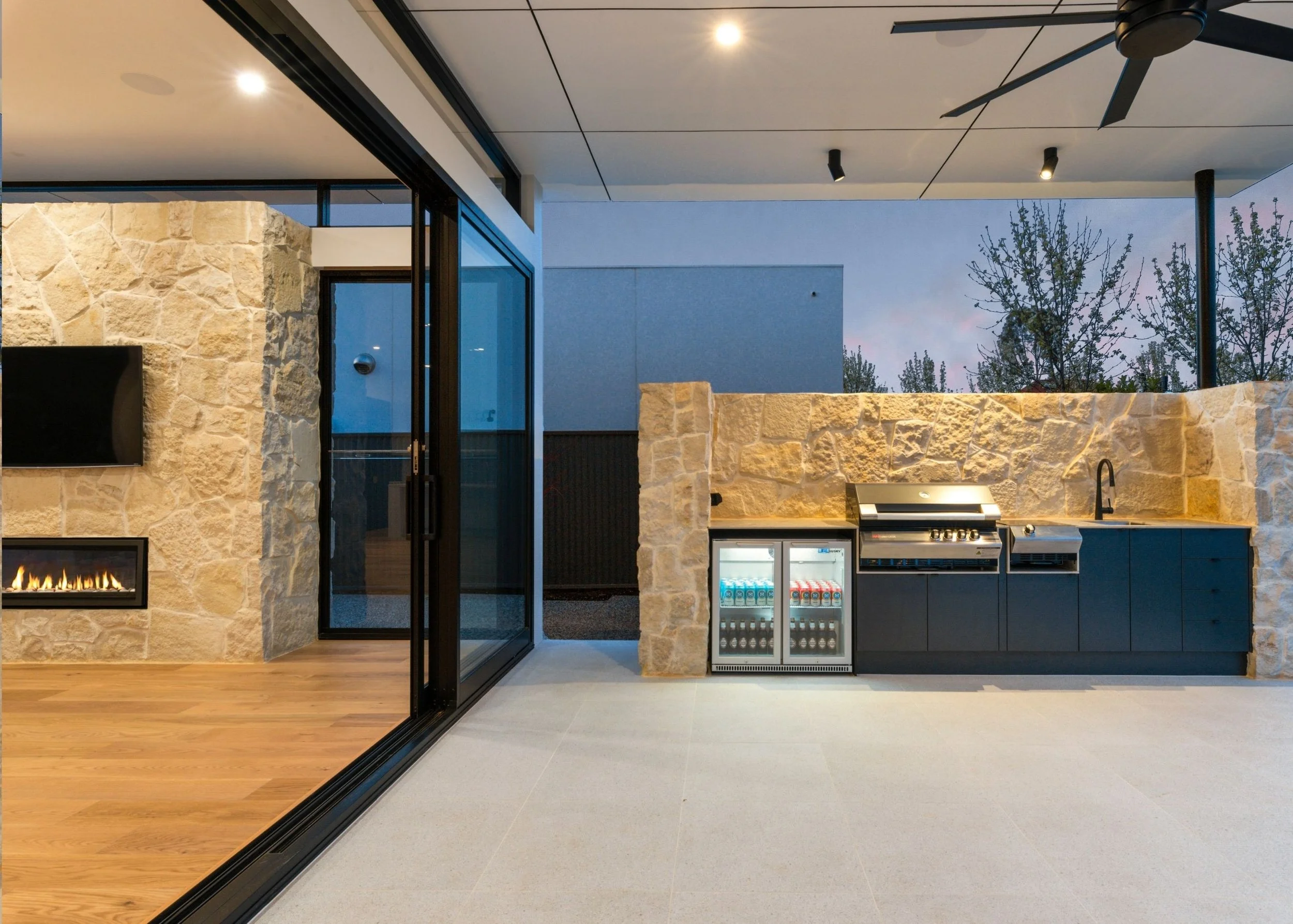 Outdoor kitchen with stone walls, built-in fireplace, mini refrigerator, and barbecue grill, with a glass sliding door leading to an indoor space and trees visible in the background.