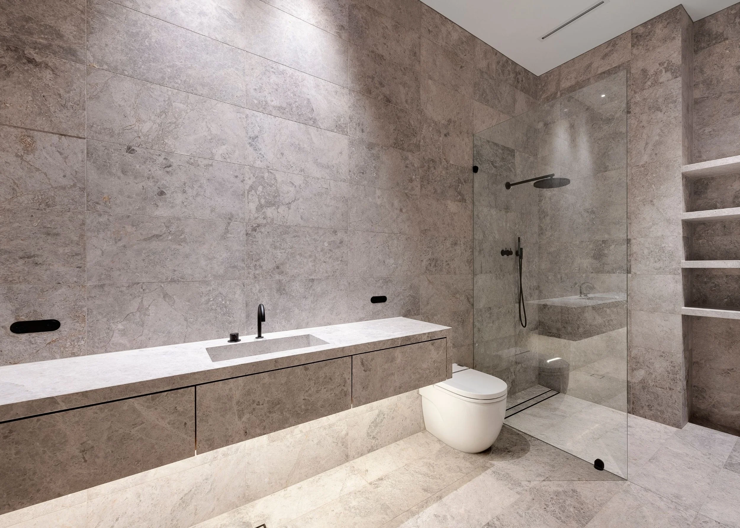 Modern bathroom with large beige marble tiles on walls and floor, a walk-in shower with glass panels, a rain shower head, a black handheld shower, a white toilet, and a beige vanity with a black faucet.