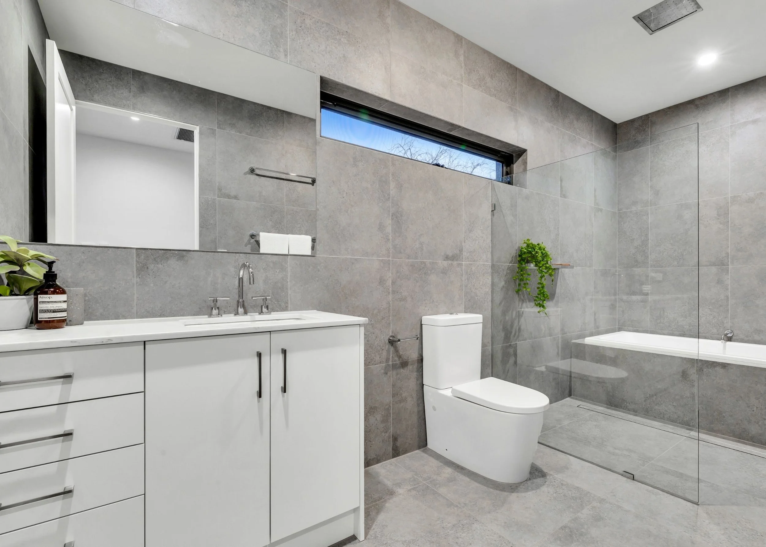 Modern bathroom with gray tiled walls and floor, white vanity with black handles, large mirror, toilet, bathtub, small window with trees outside, and green plants.