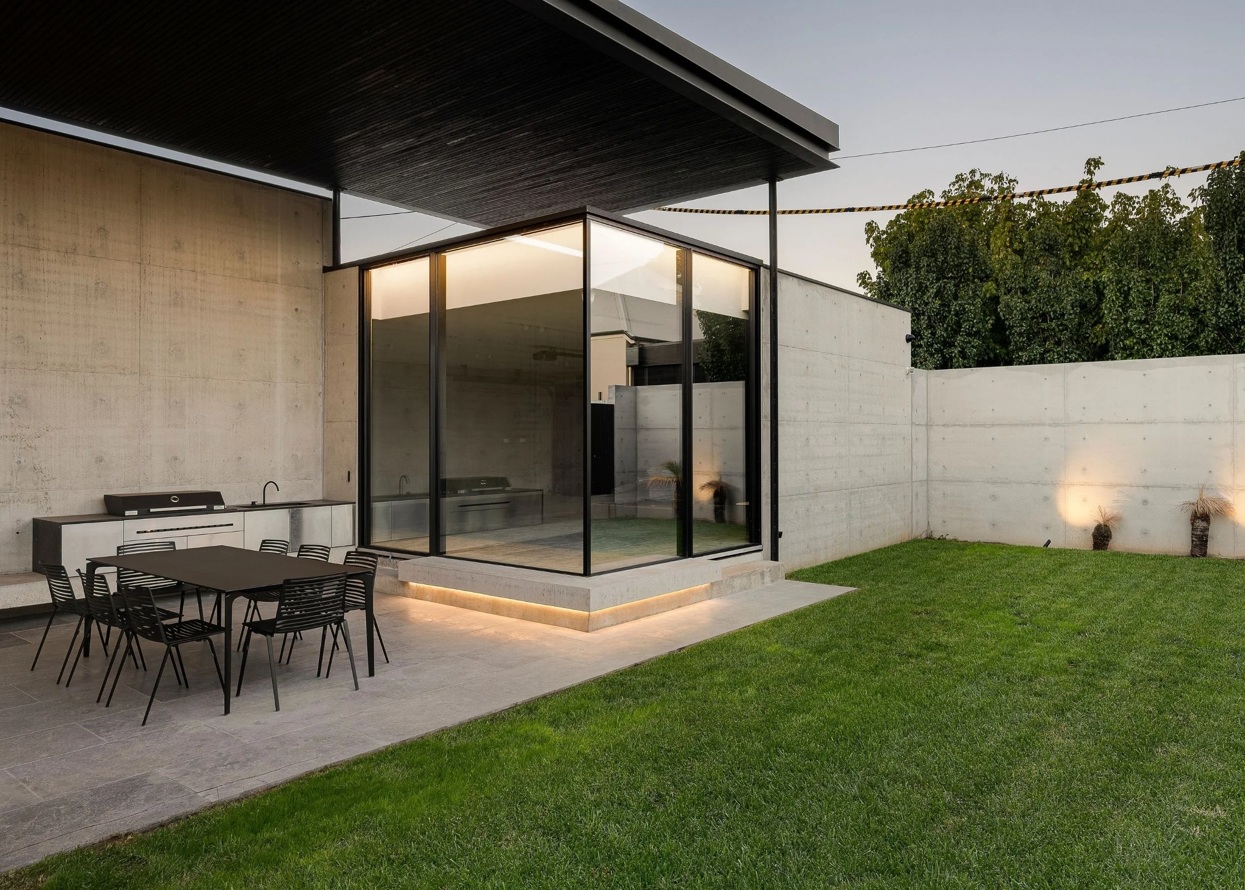 Modern house with a covered outdoor patio, glass walls, a concrete wall, a green lawn, and outdoor furniture.
