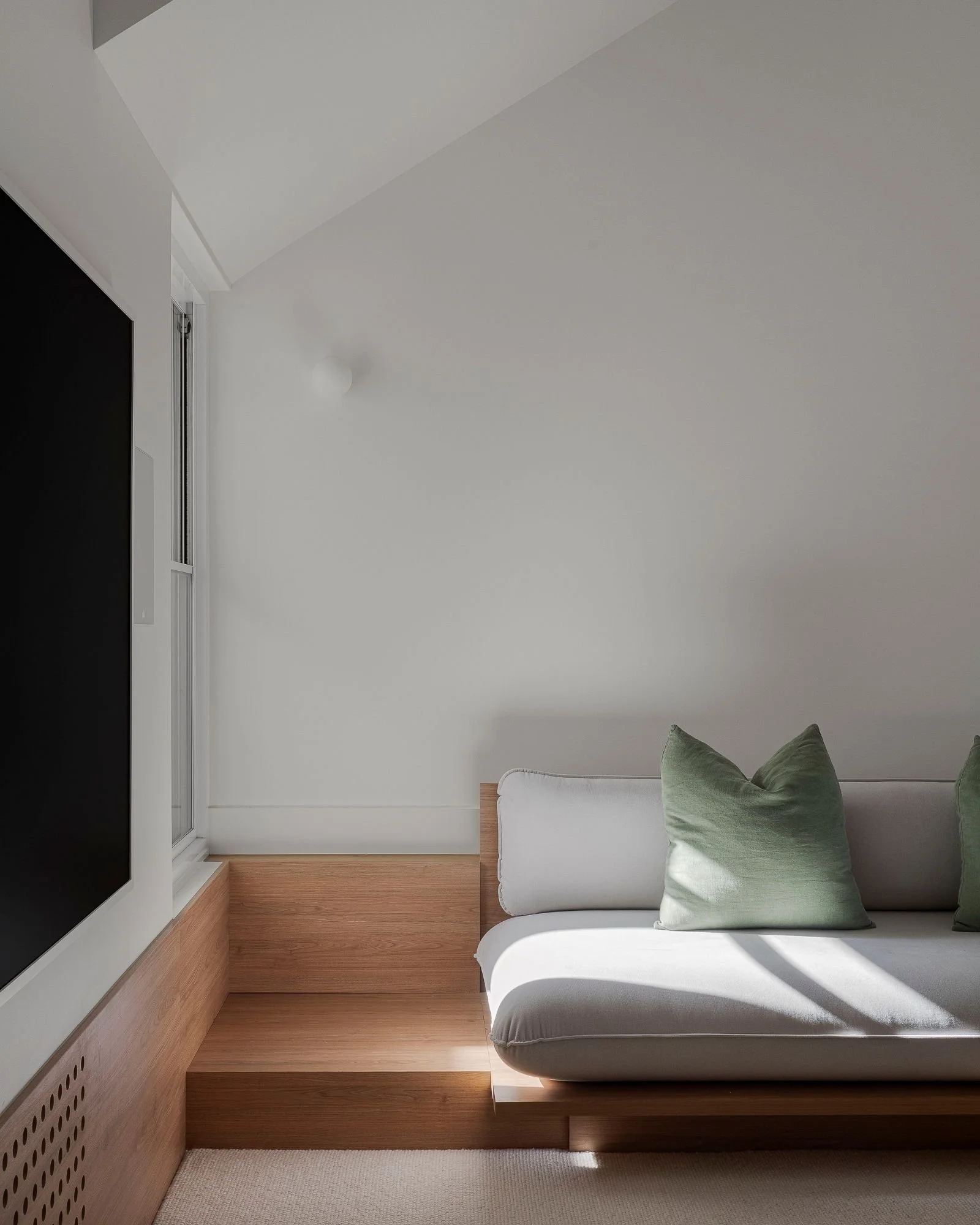 A minimalist interior corner with a white sofa, green cushions, wooden flooring, and a white wall with a window and wall-mounted light