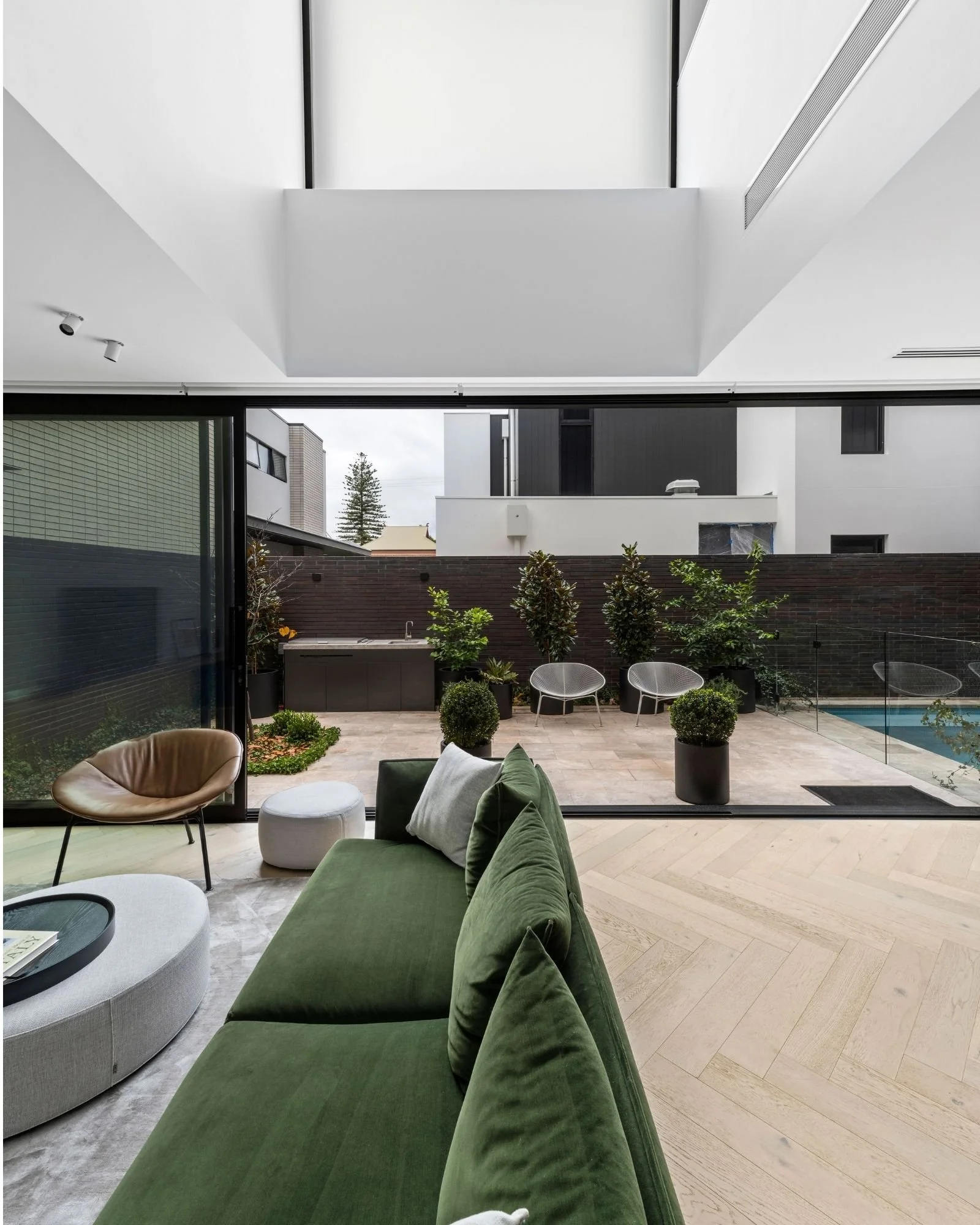 Modern indoor living area with large glass sliding door opening to outdoor patio with chairs, plants, and swimming pool.
