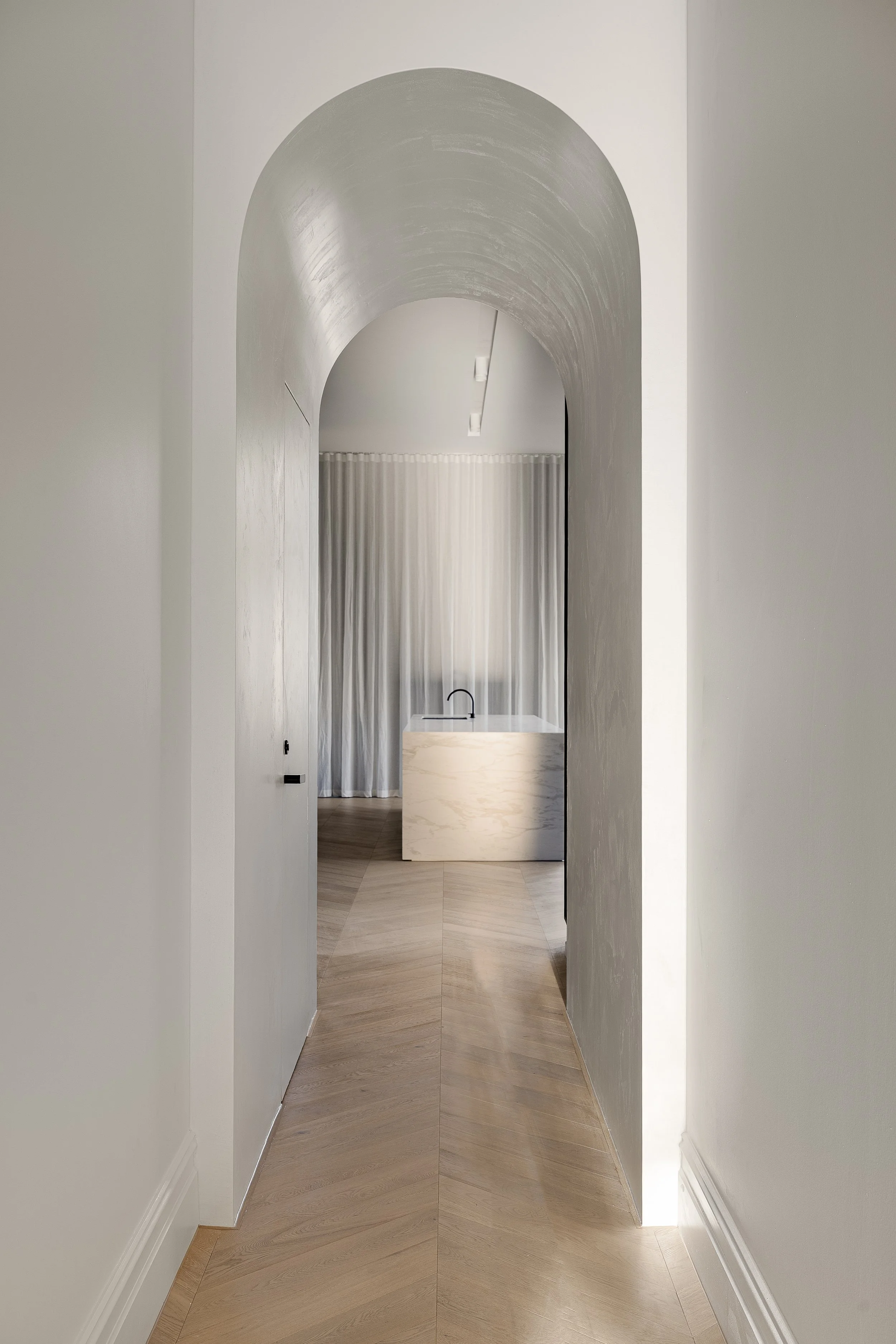 A minimalist hallway with wooden chevron-patterned flooring, white walls, an arched ceiling, and a partially visible doorway in the distance with sheer curtains and a black faucet or handle.