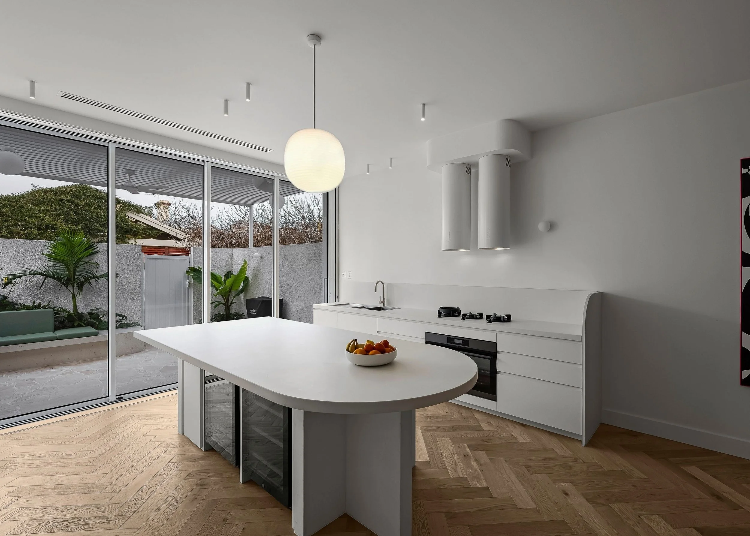 Modern white kitchen with an island, light wood floor, and large sliding glass doors opening to an outdoor patio with plants and trees.