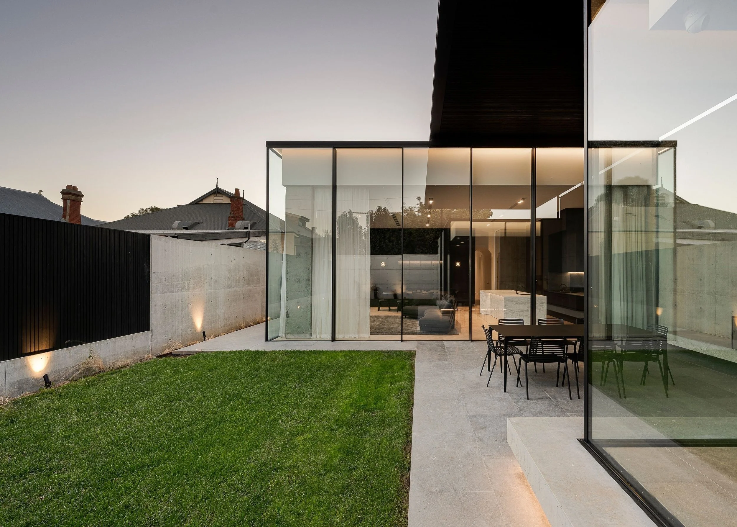 Modern house with floor-to-ceiling glass windows, outdoor patio with black dining table and chairs, adjacent green lawn, and surrounding low concrete wall.