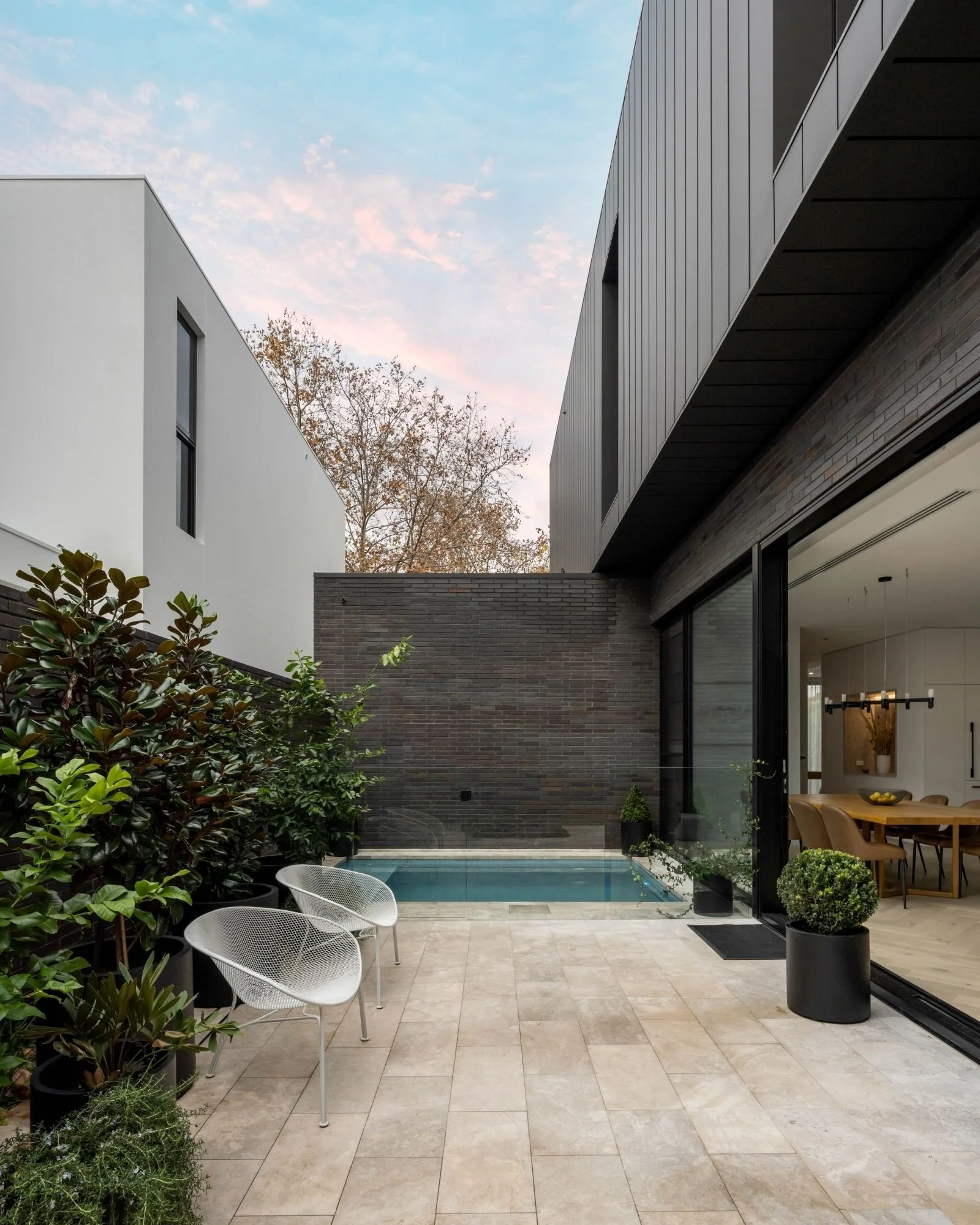 Modern house with a backyard patio, small swimming pool, outdoor furniture, potted plants, and large sliding glass doors, with a dark brick wall and a neighboring white house in the background.