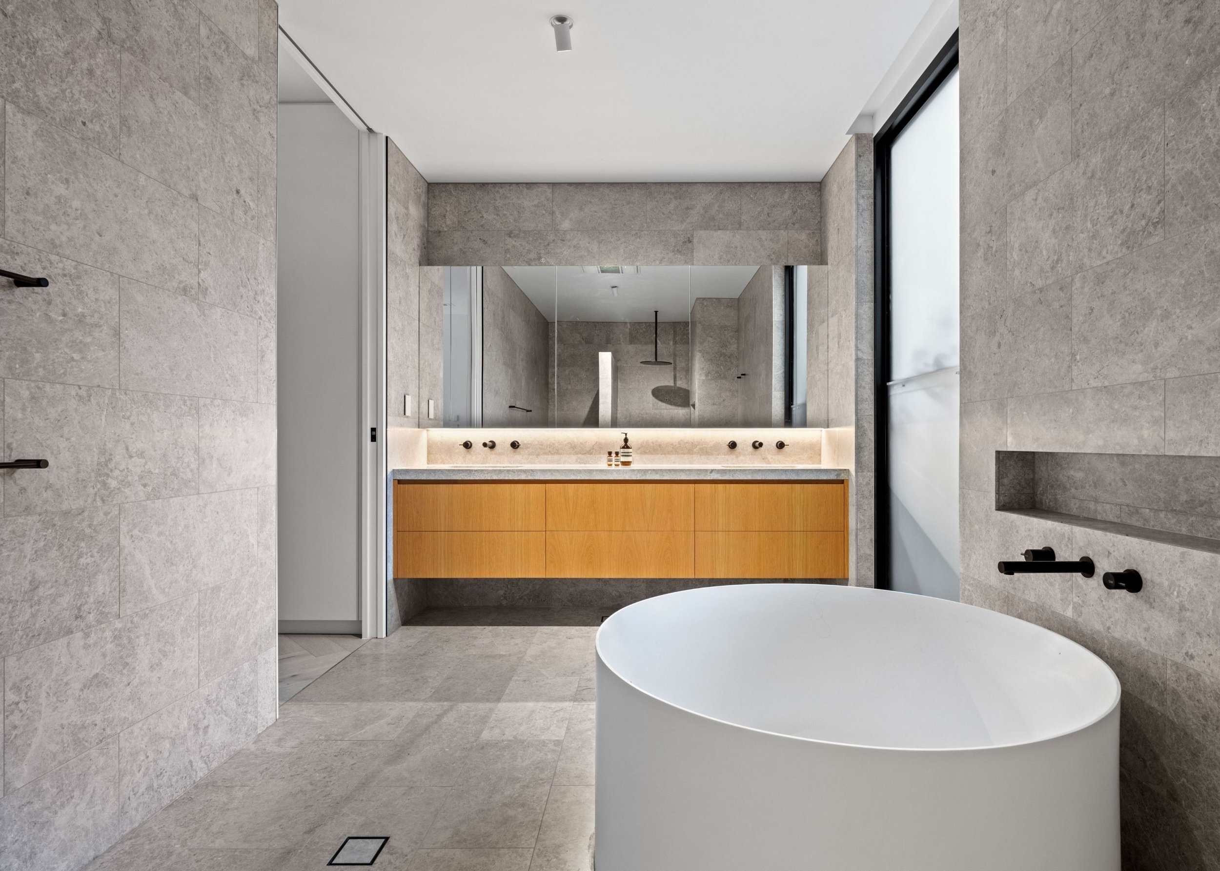 Modern minimalist bathroom with a freestanding bathtub, large mirror, wooden vanity, stone tiles, and large windows.