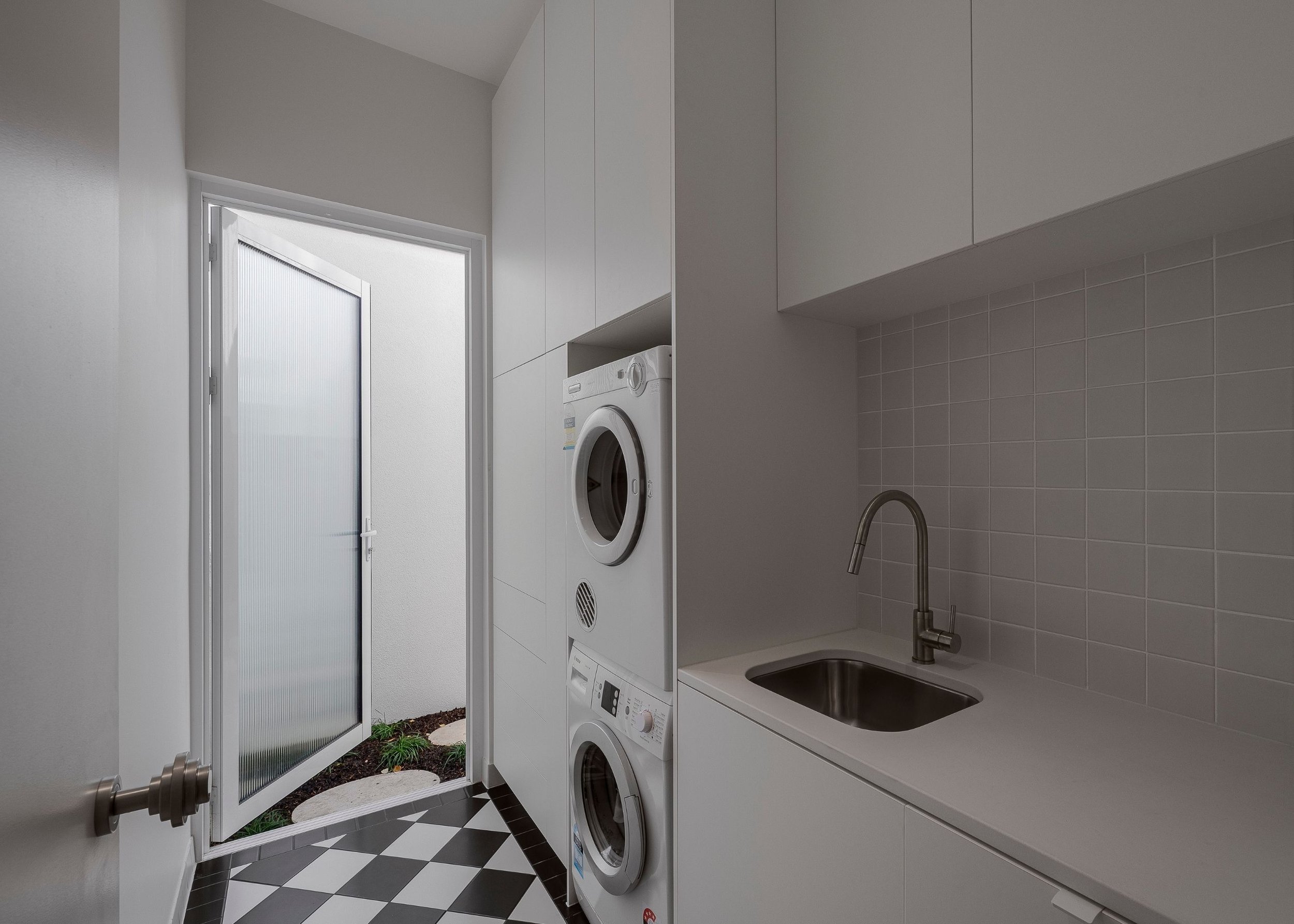 Laundry room with stacked washer and dryer, white cabinets, sink, tiled wall, door leading outside, and black-and-white checkered floor.