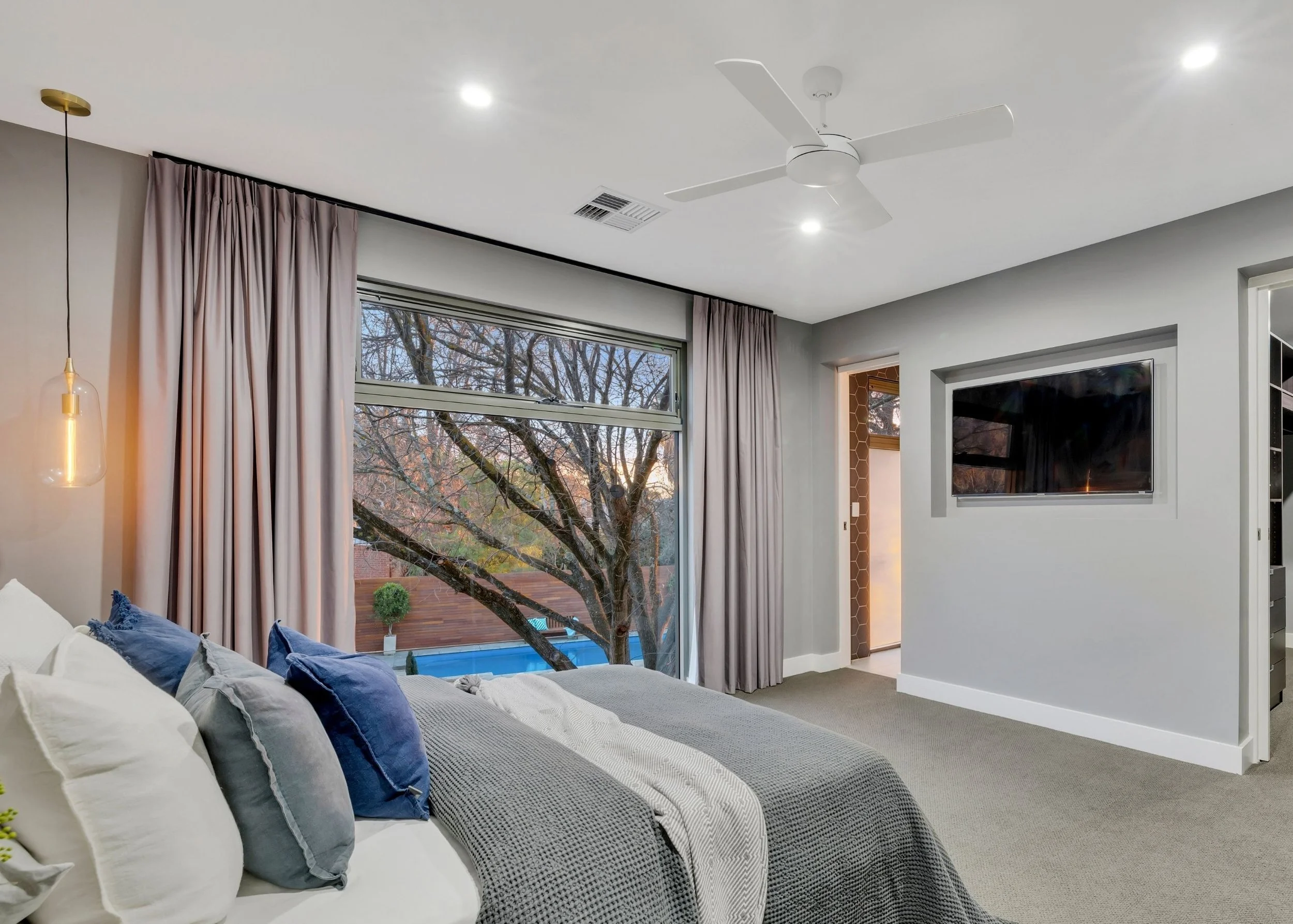 Interior view of a modern living room with a large window showing a tree outside. The ceiling has a white ceiling fan and recessed lighting. There are curtains on the window and a sofa with blue and white cushions.