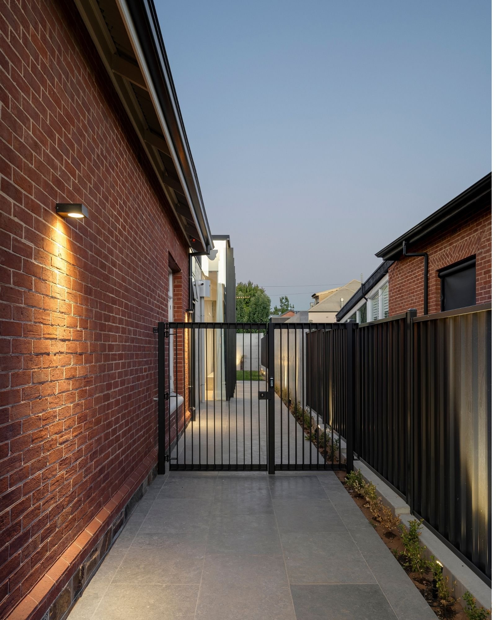 Outdoor alleyway with brick wall on the left, black metal gate in the center, black fence on the right, small plants along the fence, and modern house exterior with windows and roof in the background, under a clear blue sky.