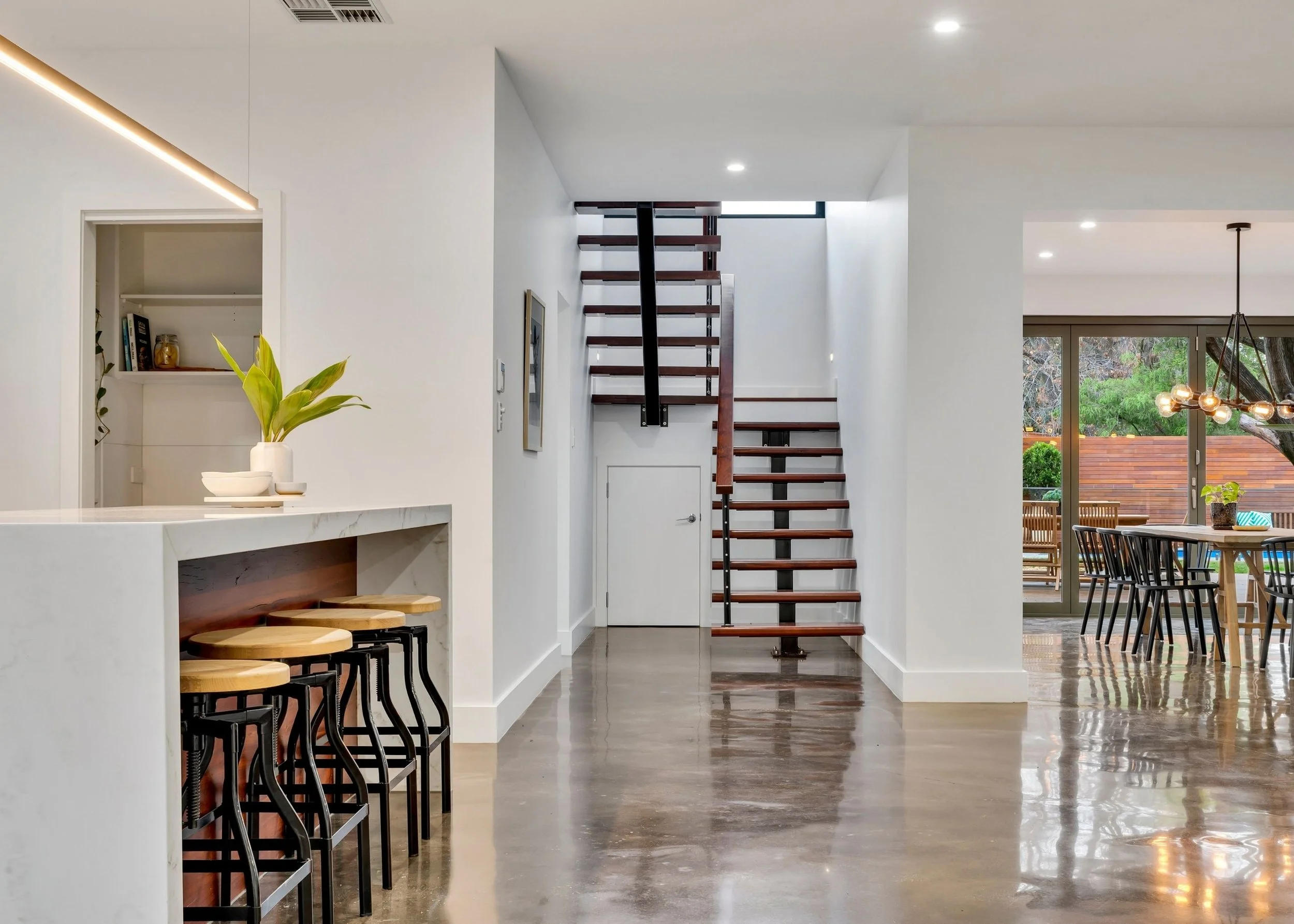 Interior view of a modern home with a staircase, dining area, and a kitchen island with barstools, featuring white walls, wooden floors, and a sliding glass door leading to an outdoor patio.