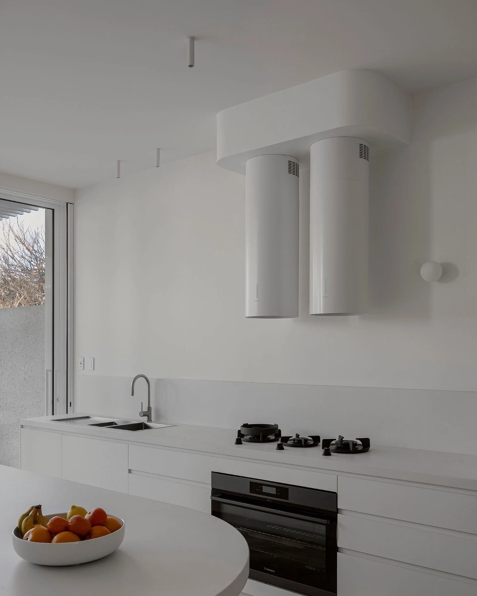 Modern white kitchen with built-in oven, stovetop, white cabinets, and two suspended cylindrical range hoods; window showing outdoor trees; bowl of fruit on white round table.