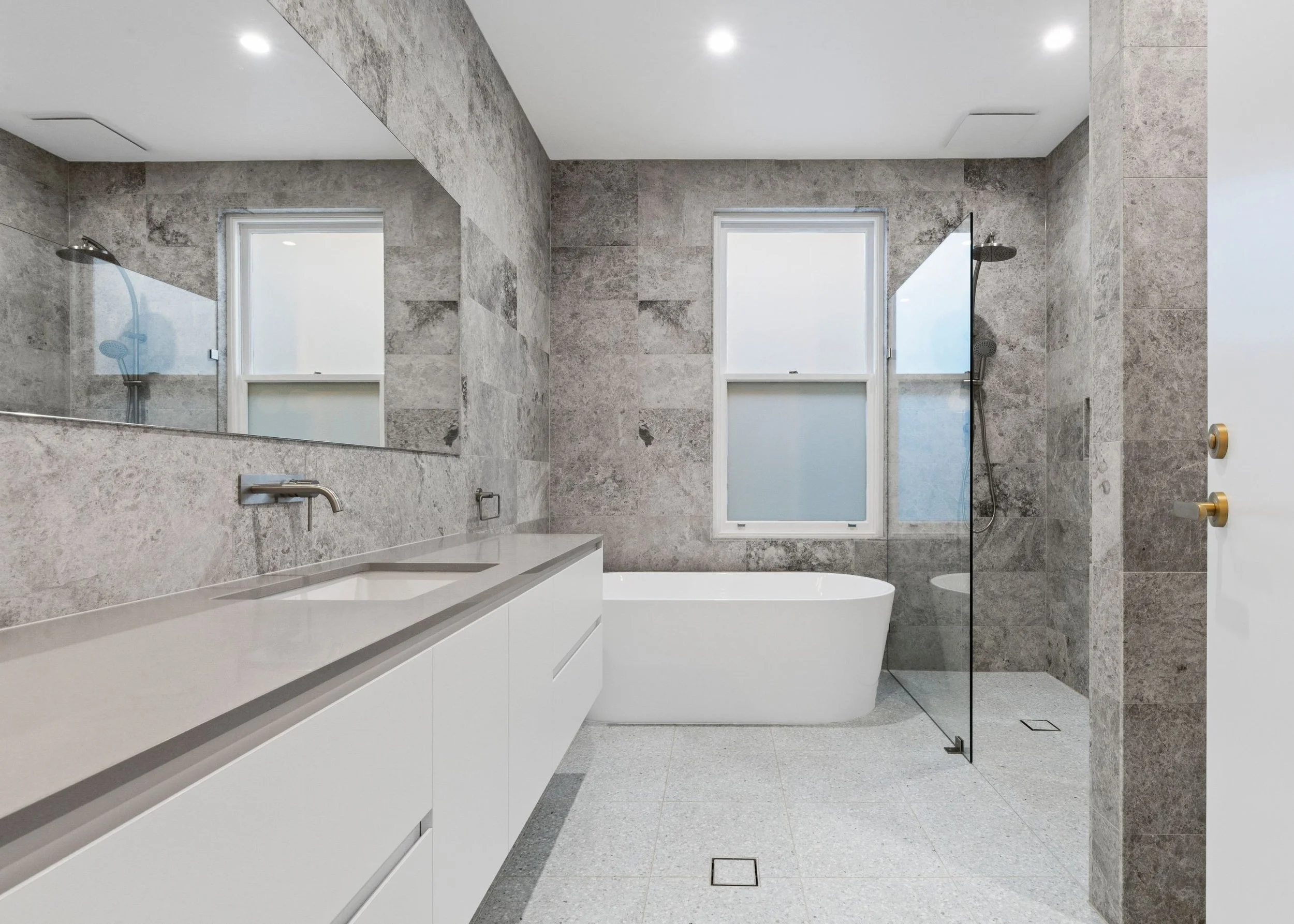 Modern bathroom with gray stone walls and floors, a bathtub, two large windows, a glass shower door, and white cabinetry.