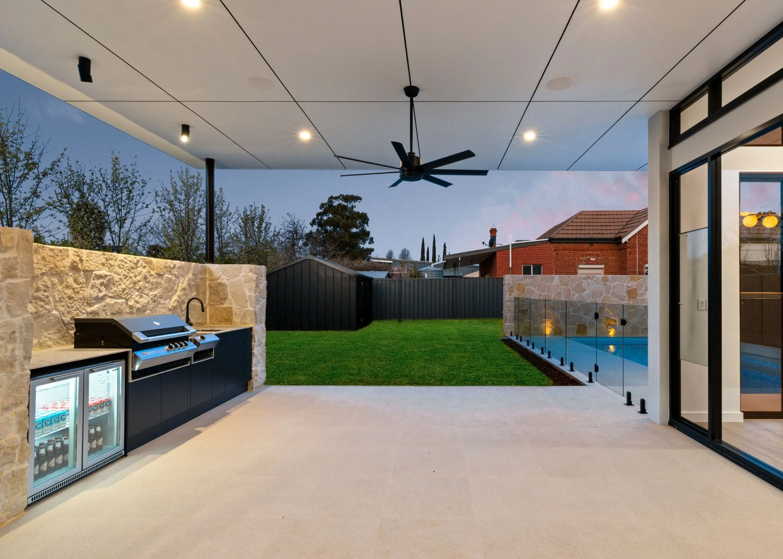 Modern house patio with outdoor kitchen, ceiling fan, and a view of a backyard with a pool, fence, and houses beyond at dusk.