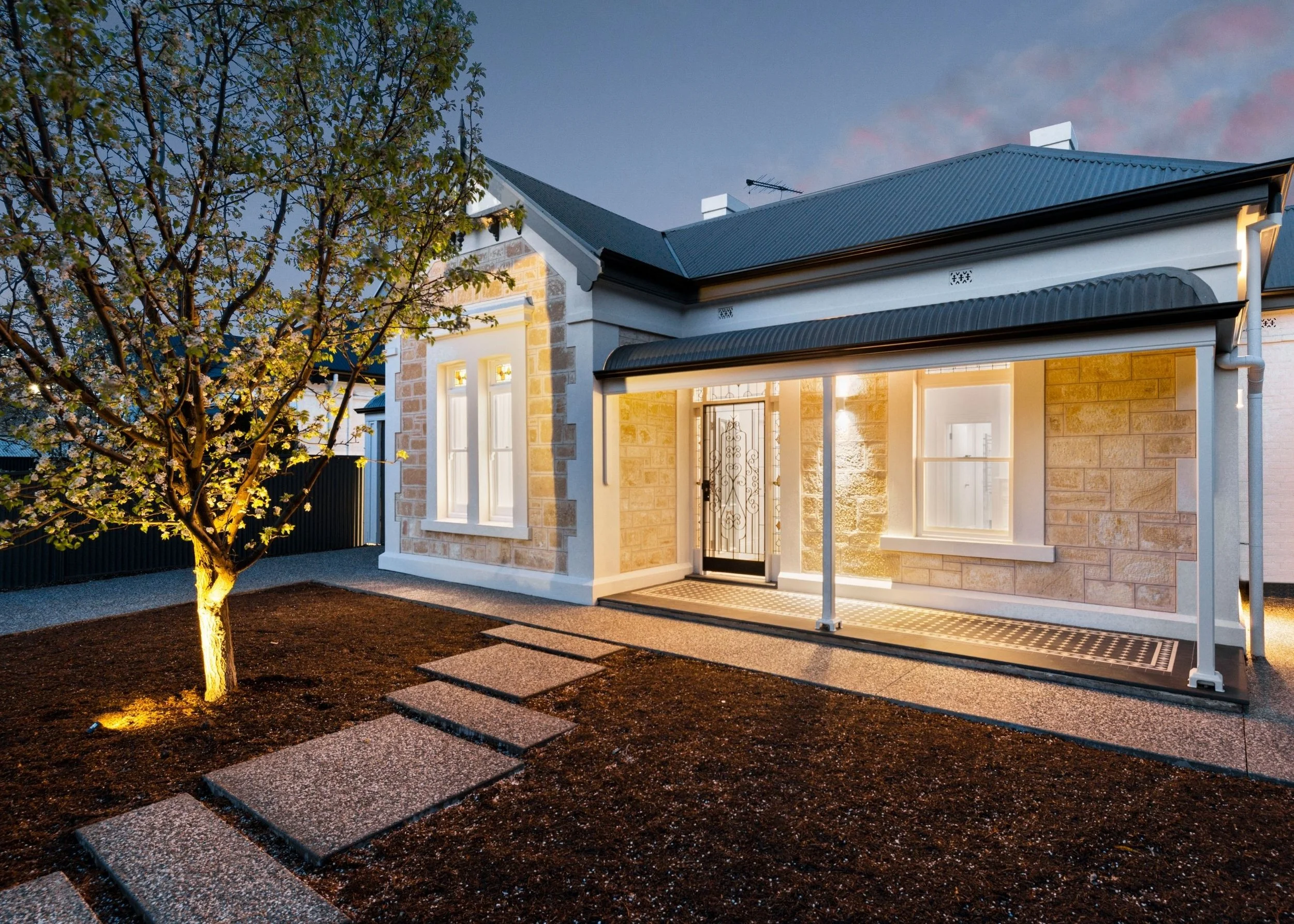 A modern house with a black metal roof, stone and white exterior walls, illuminated windows, a sidewalk, and a flowering tree outside, taken during dusk.