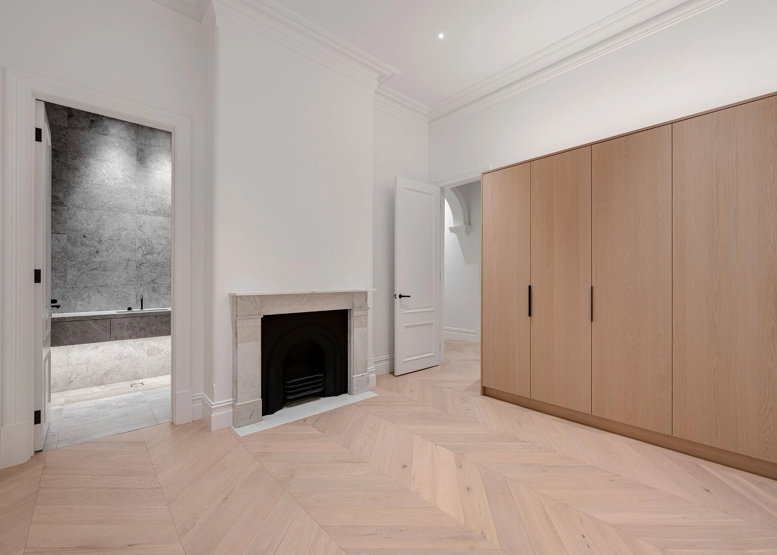 A modern interior room with a white ceiling, crown molding, and a small, round recessed light. The room features light-colored wooden flooring arranged in a herringbone pattern, a light wood door, a fireplace with a white marble mantel, and large woo
