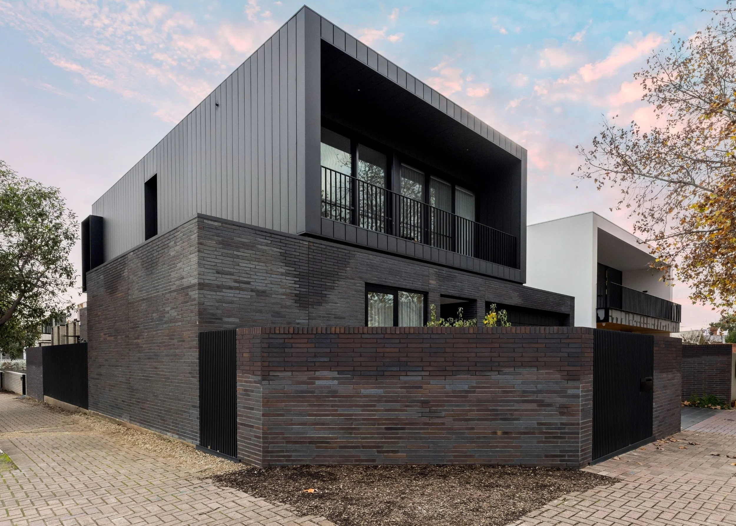 Modern multi-story house with a brick lower level and gray upper level, featuring large windows and a balcony, surrounded by trees and a paved sidewalk, during early evening.
