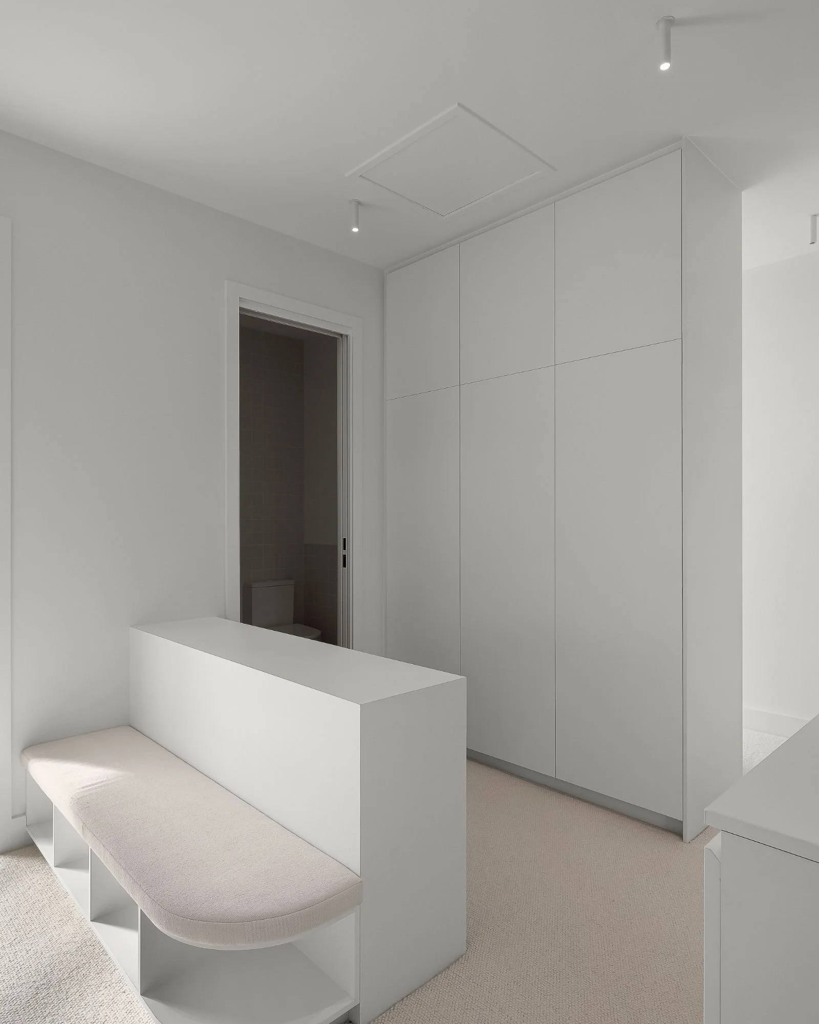 Minimalist white room with a built-in white wardrobe, a white couch, and a small section of a white table. There is an open door leading to a small bathroom with tiled walls, and a ceiling with small spotlights.