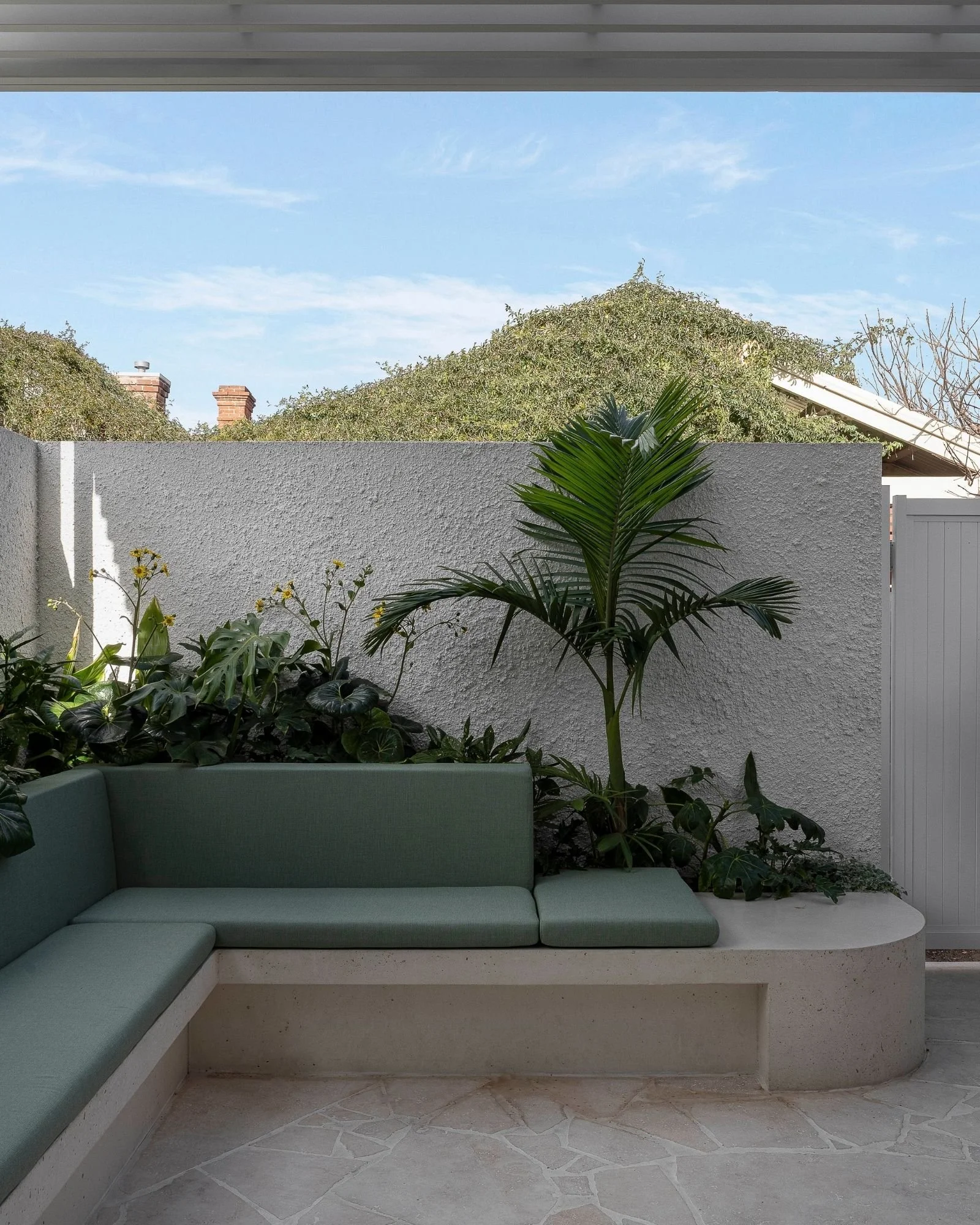 A backyard patio with a green cushioned built-in bench, surrounded by potted plants and shrubs, enclosed by a concrete wall, with trees and a house roof visible above the wall.