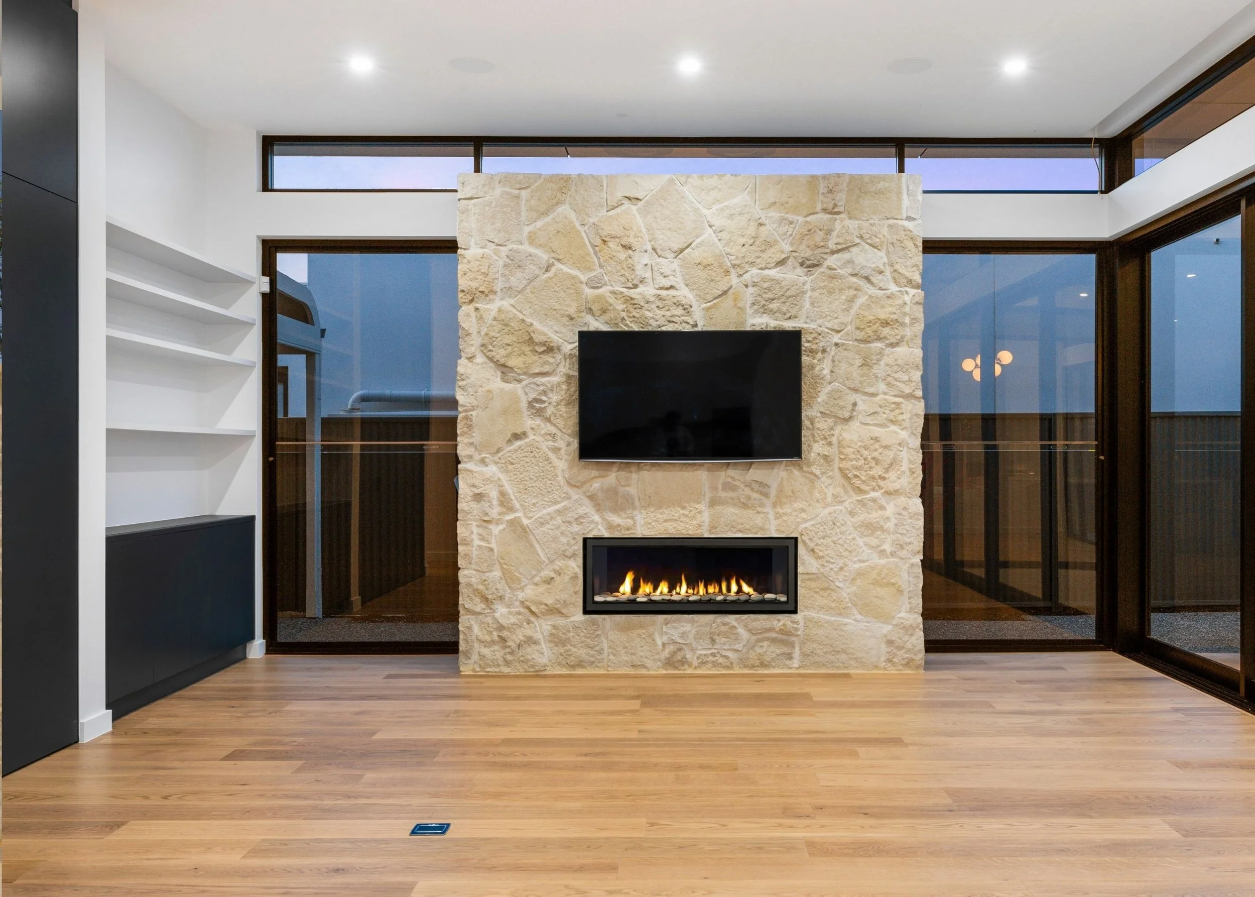 Interior of a modern room with a stone fireplace, mounted TV, large windows, hardwood flooring, and minimal decor.