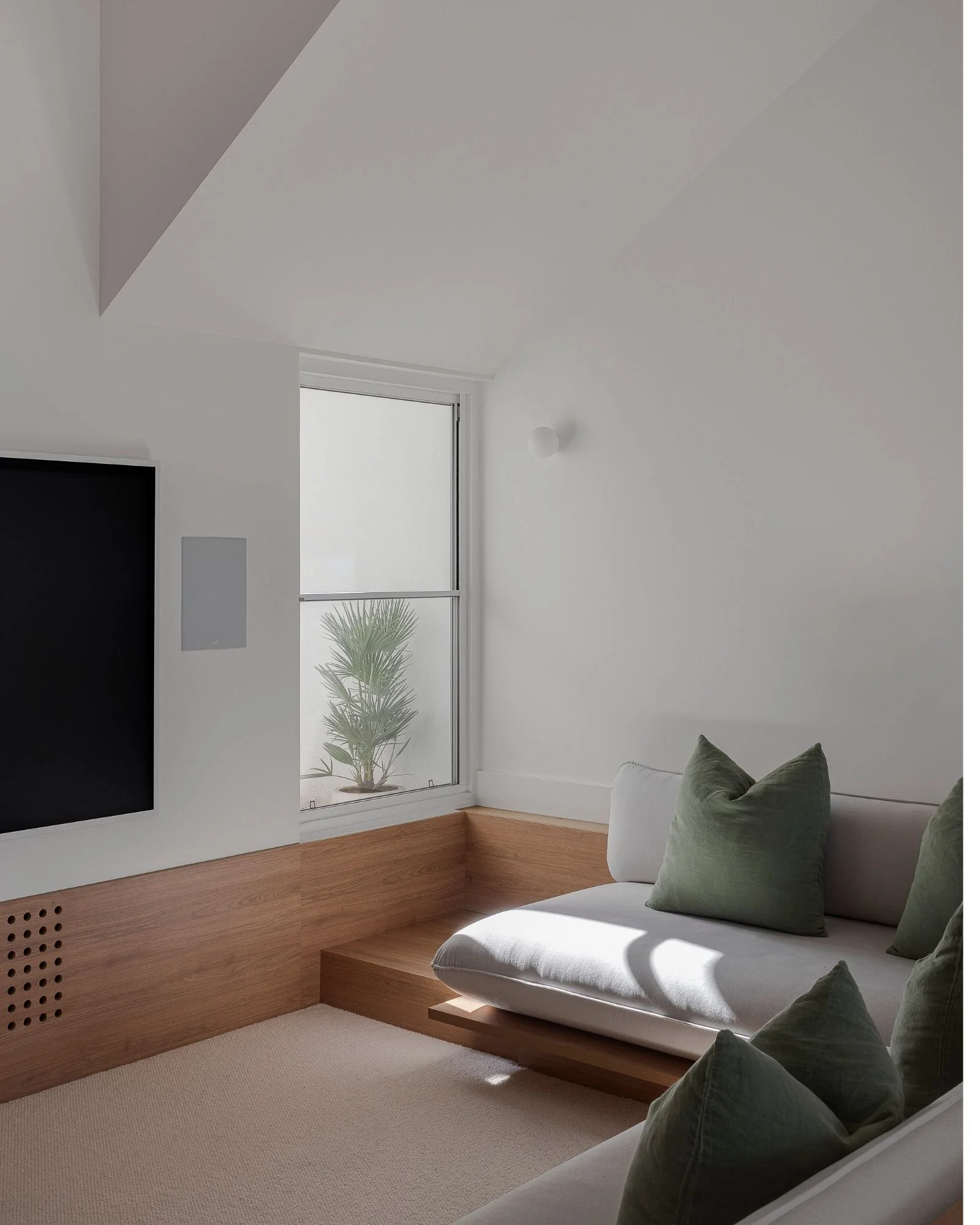 A modern, minimalist living room with white walls, a window with a view of a plant outside, a white sofa with green pillows, a wooden baseboard, and a light carpet.