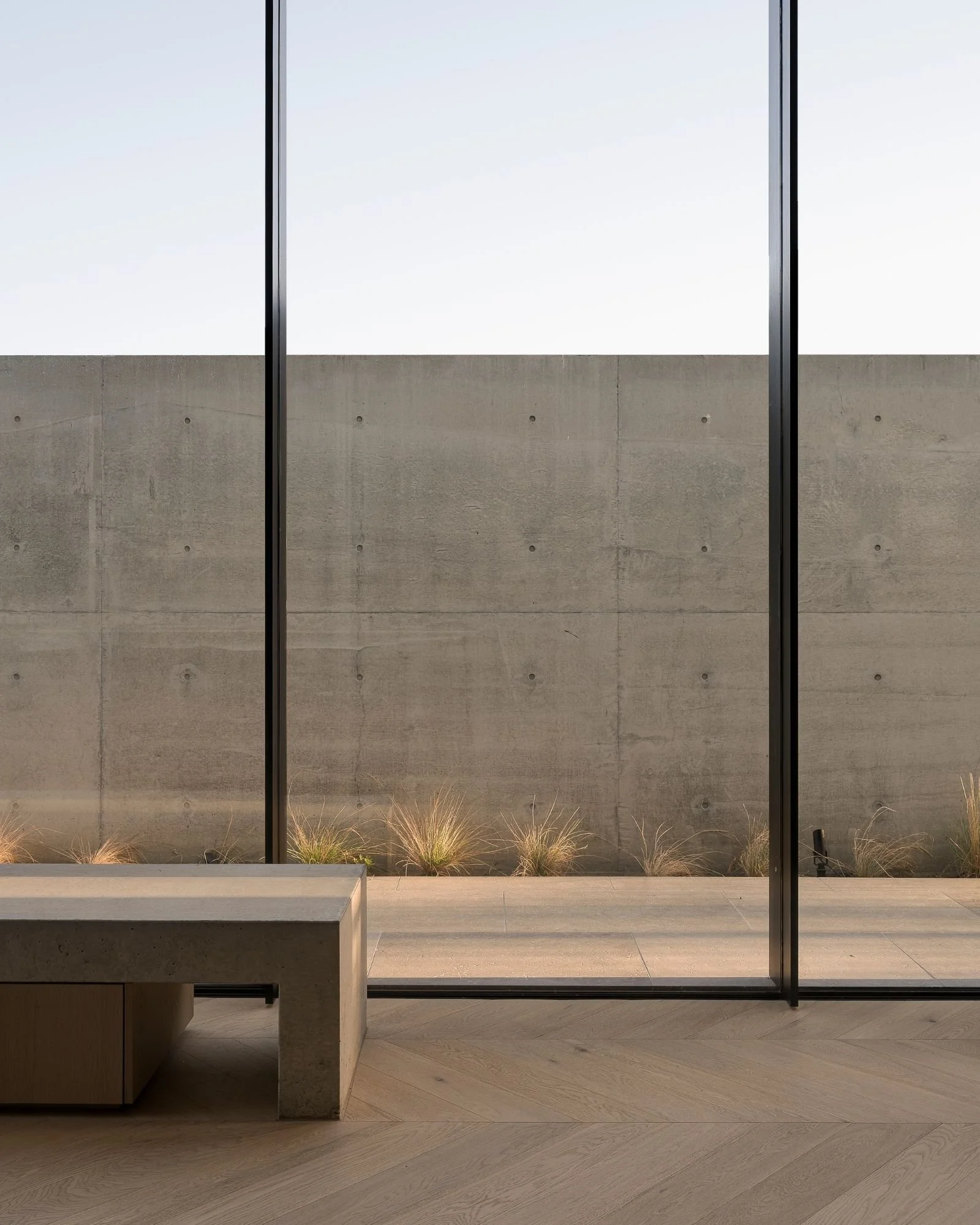 Indoor space with a large window showing a view of a concrete wall and some plants outside, modern minimalist decor, light wood flooring.