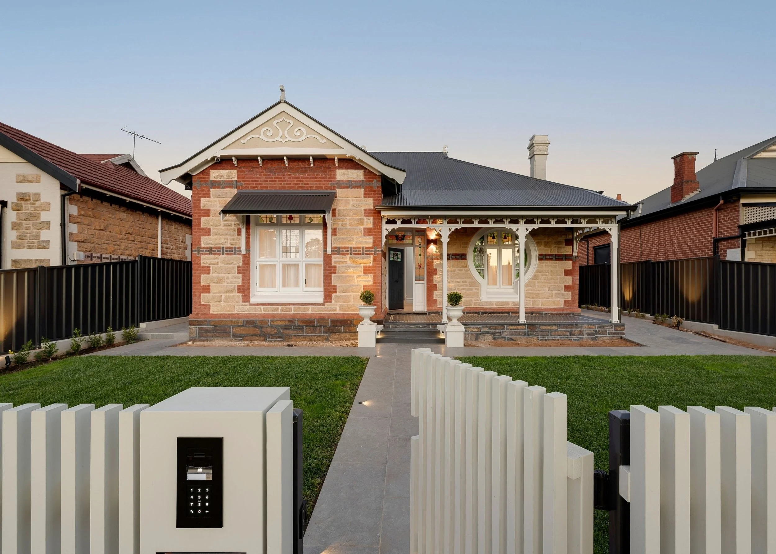 A charming house with red brick and beige stone exterior, black roof, and decorative white trim, featuring a front porch with white railings and two white planters, surrounded by a white picket fence, with modern security keypad at the gate, set agai