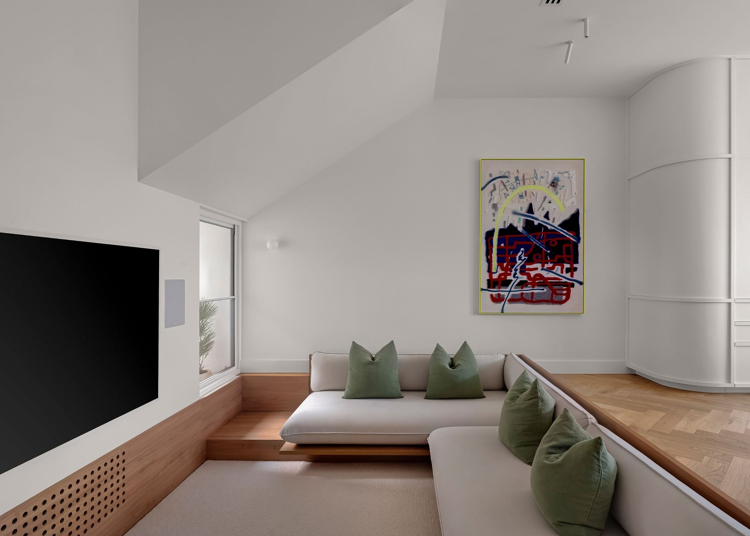 Modern living room with white walls, wooden accents, a built-in seating area with green pillows, a large white couch, abstract artwork on the wall, and windows letting in natural light.