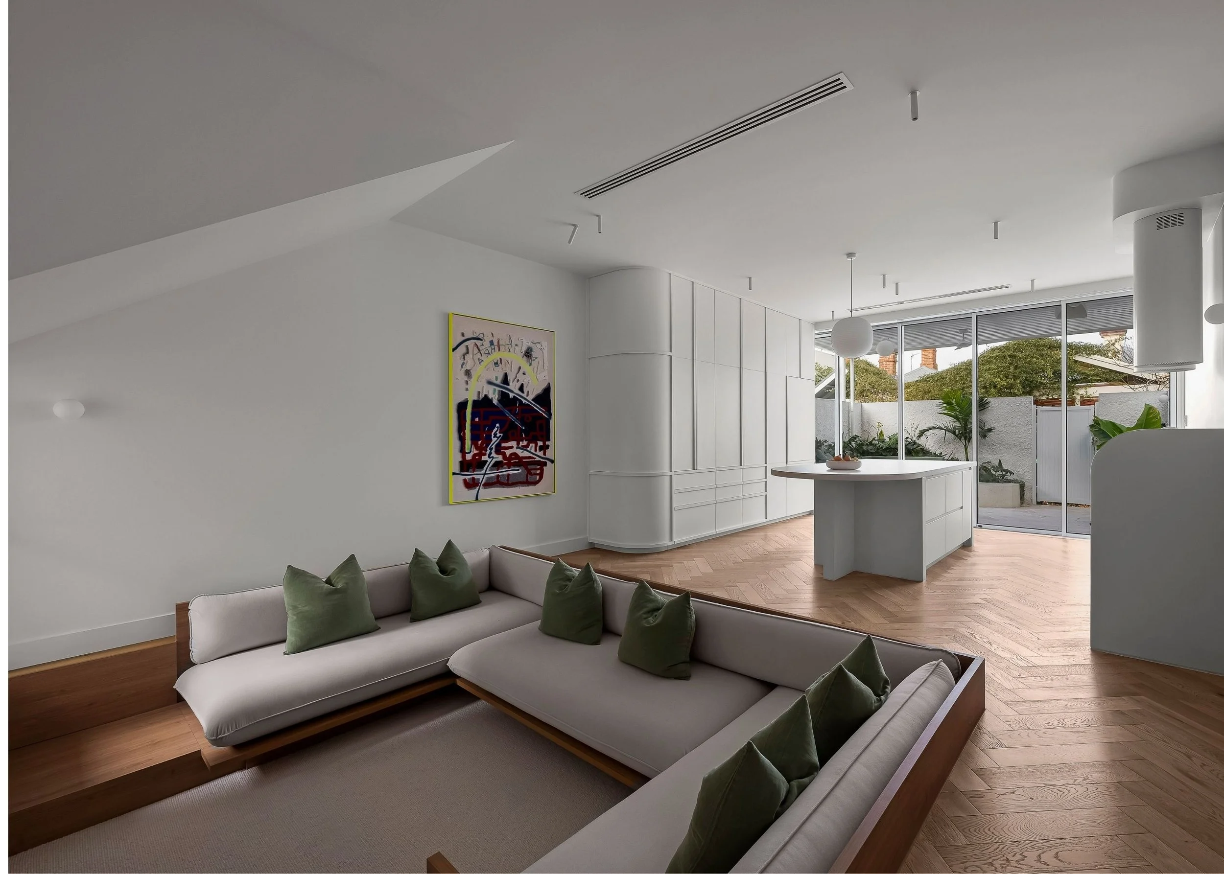 Modern living room with white walls, wooden floor, white sofa with green pillows, abstract art on wall, and large glass sliding doors leading outside to a patio with greenery.