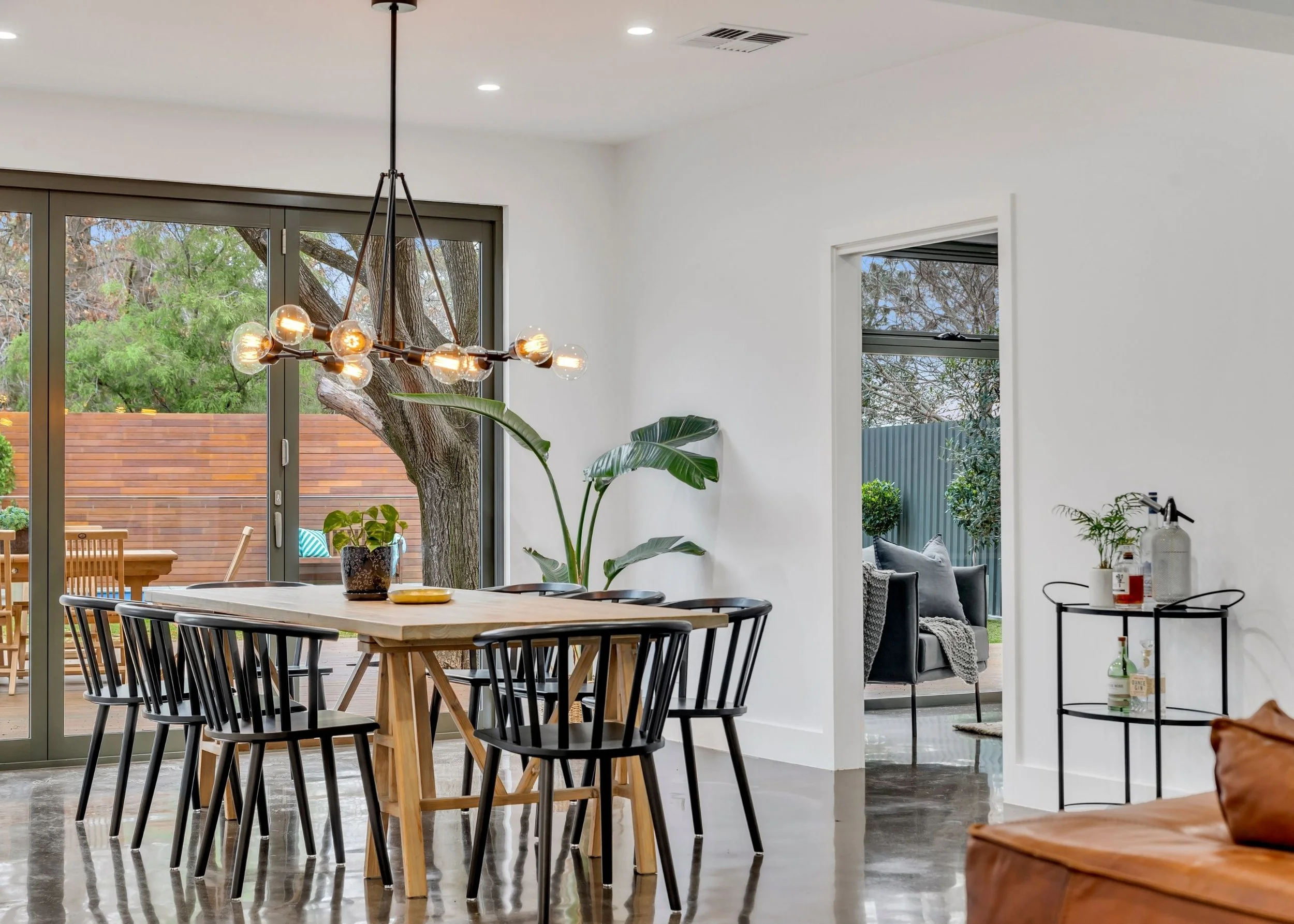 Modern dining and living area with a large wooden table and black chairs, a chandelier hanging from the ceiling, large sliding glass doors leading to an outdoor patio with trees, and a glimpse of a sitting area with a couch and decorative plants indo