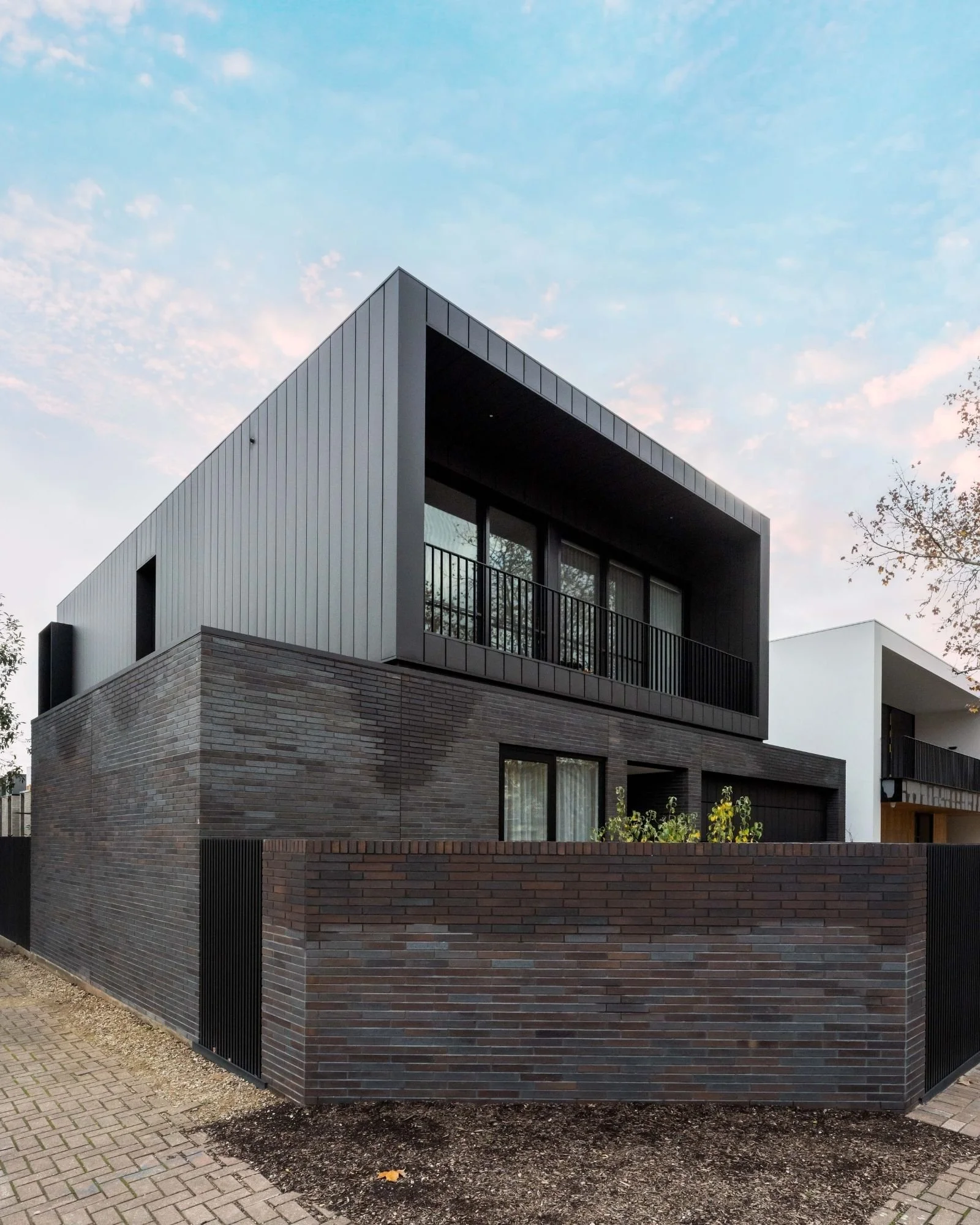 Modern two-story house with brick and metal exterior, large windows, and a balcony with black railing, surrounded by a paved walkway and garden, under a partly cloudy sky.