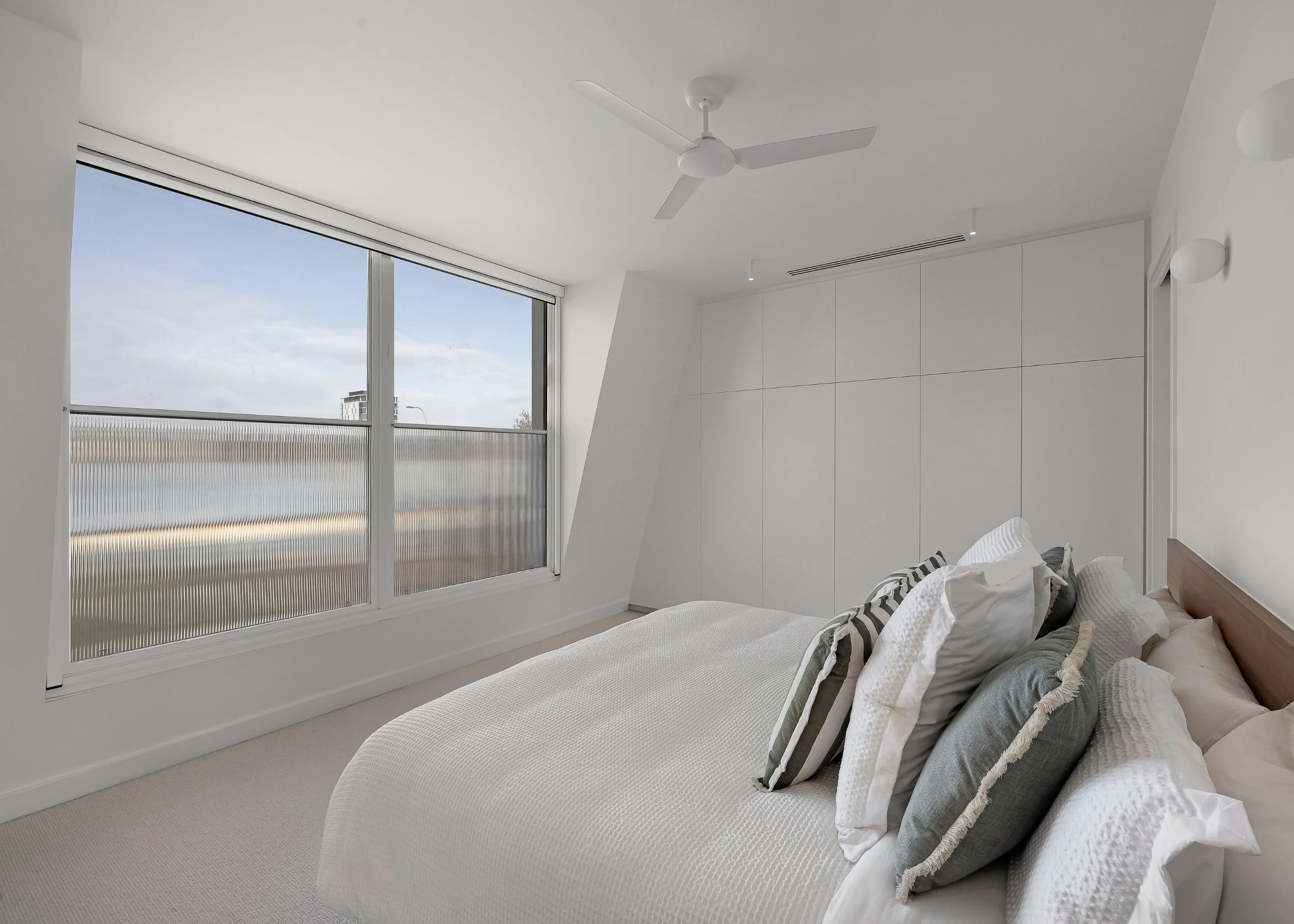 A modern bedroom with a large window, a white ceiling fan, a white built-in wardrobe, and a bed with white and striped pillows.
