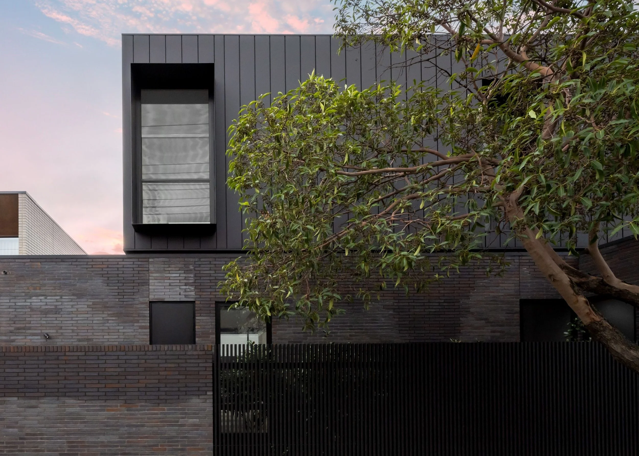 Modern building with dark gray panels and large windows, partially obscured by a large leafy tree, under a pink and blue sky at sunset.