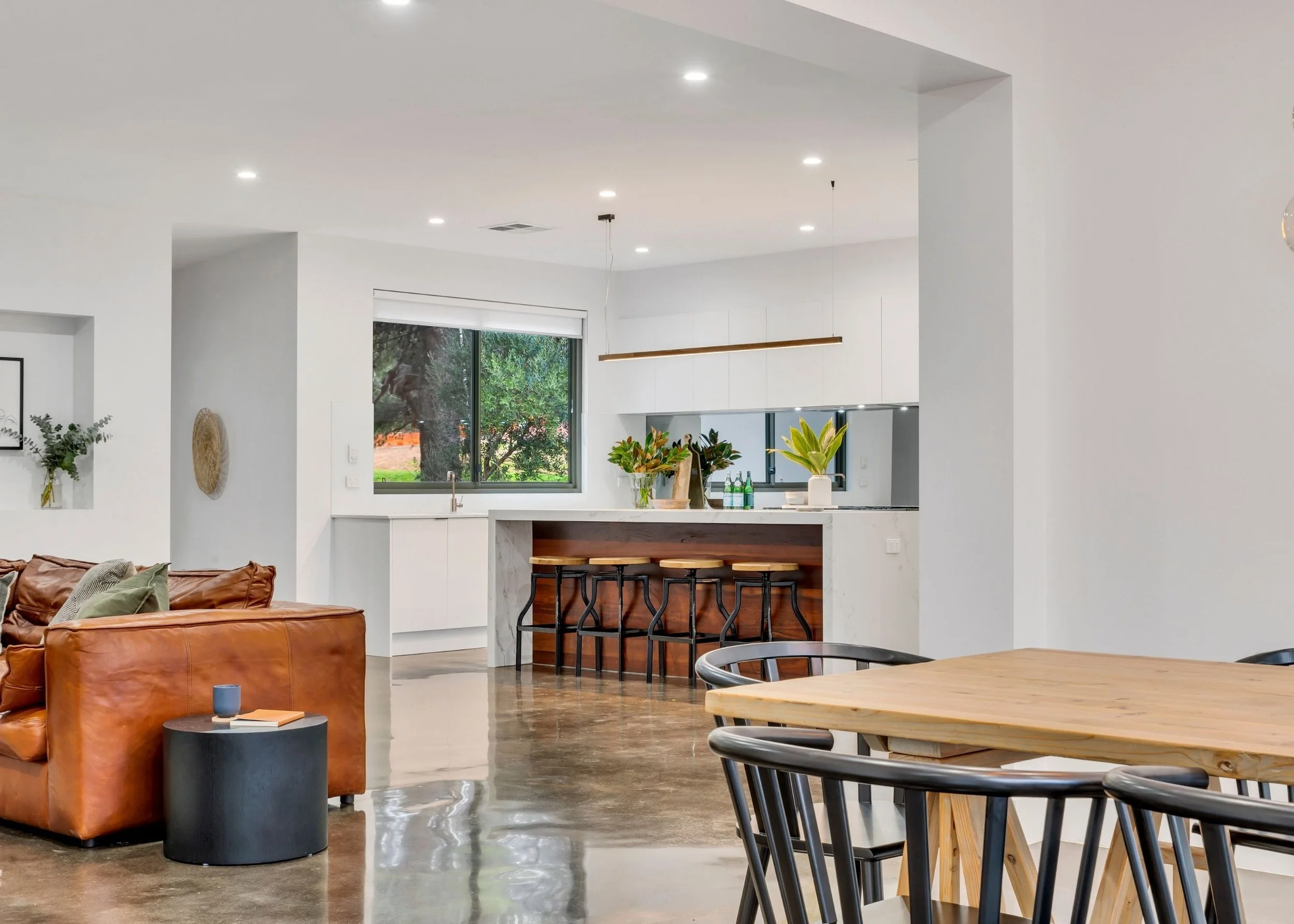 Open-concept living space featuring a kitchen with white cabinets, a window with greenery outside, a tan leather sofa, a small round black side table with a candle, a wooden dining table with black chairs, and indoor plants.