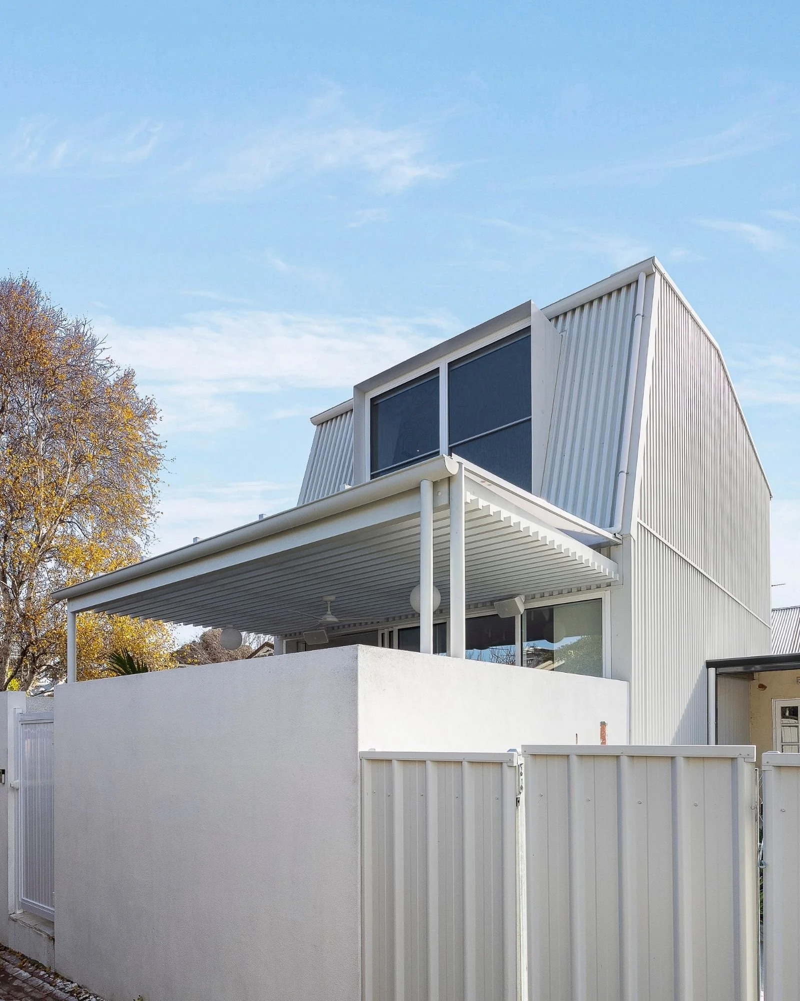 Modern white building with large windows, metal siding, balcony, and a privacy wall, under a blue sky.