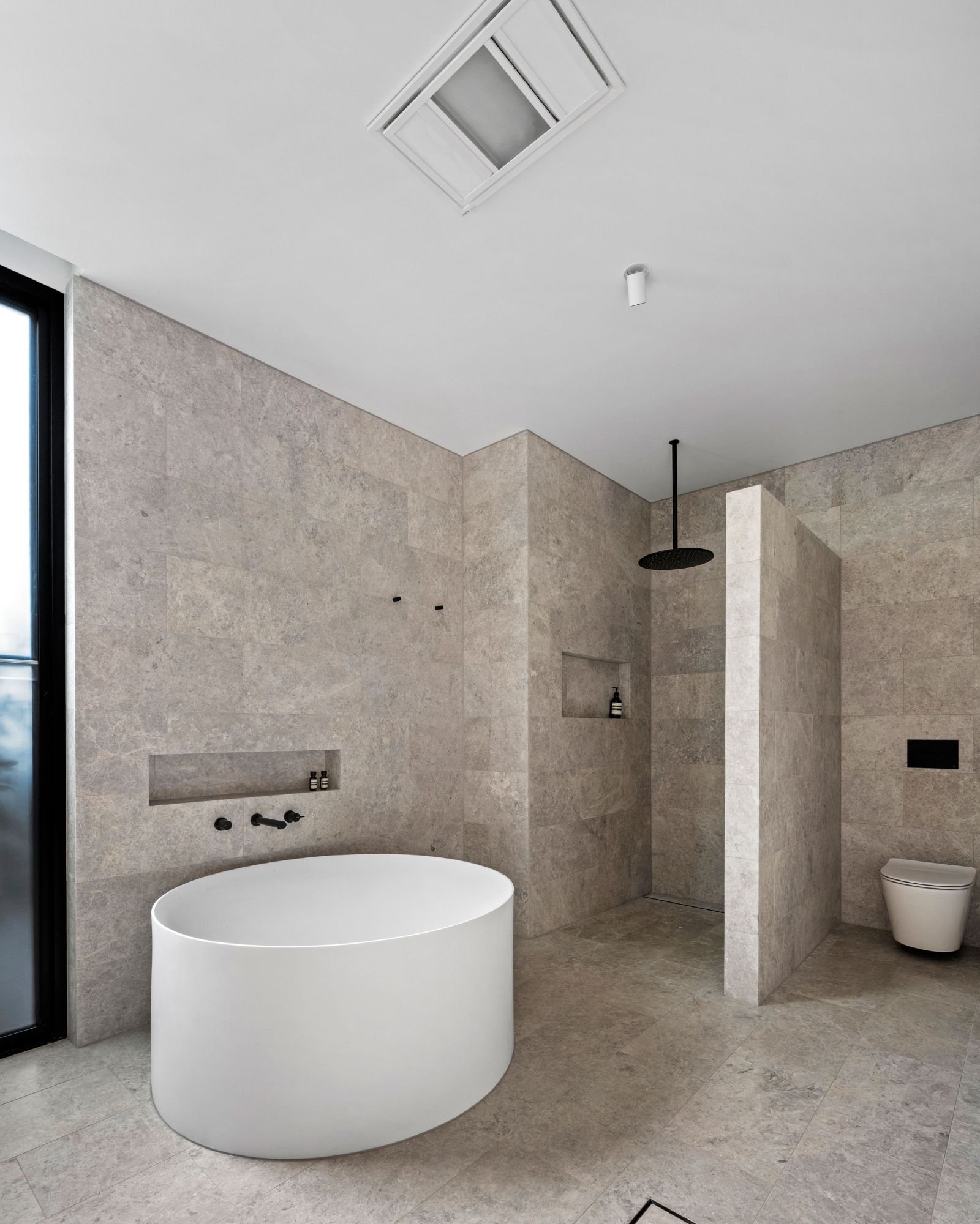 Modern bathroom with beige tile walls and floor, a freestanding white bathtub, shower area with black shower head, niche with toiletries, ceiling vent, skylight, and toilet.