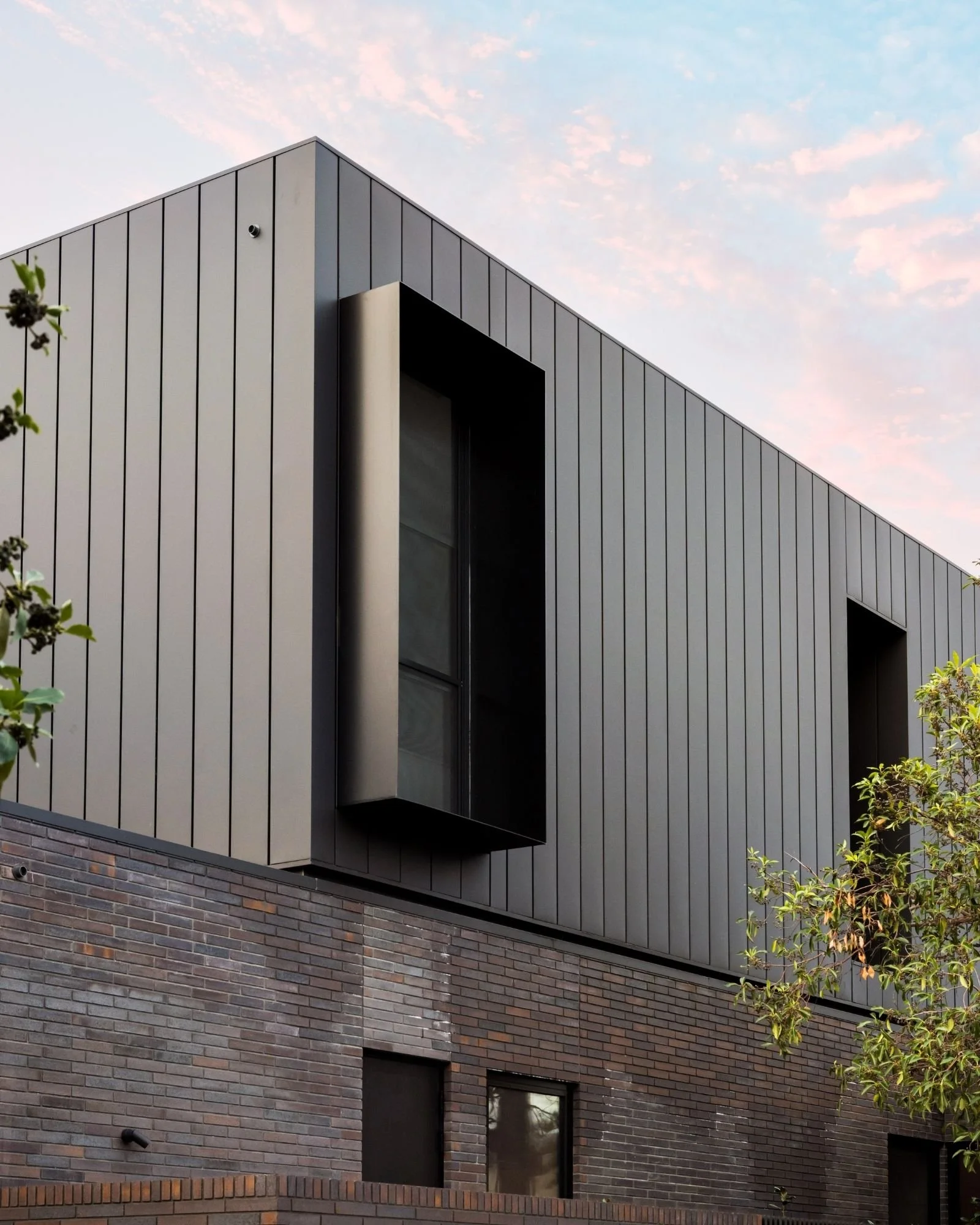 Modern building with a brick lower section and a metal upper section, featuring a small window and a black overhang, with trees and a cloudy sky in the background.