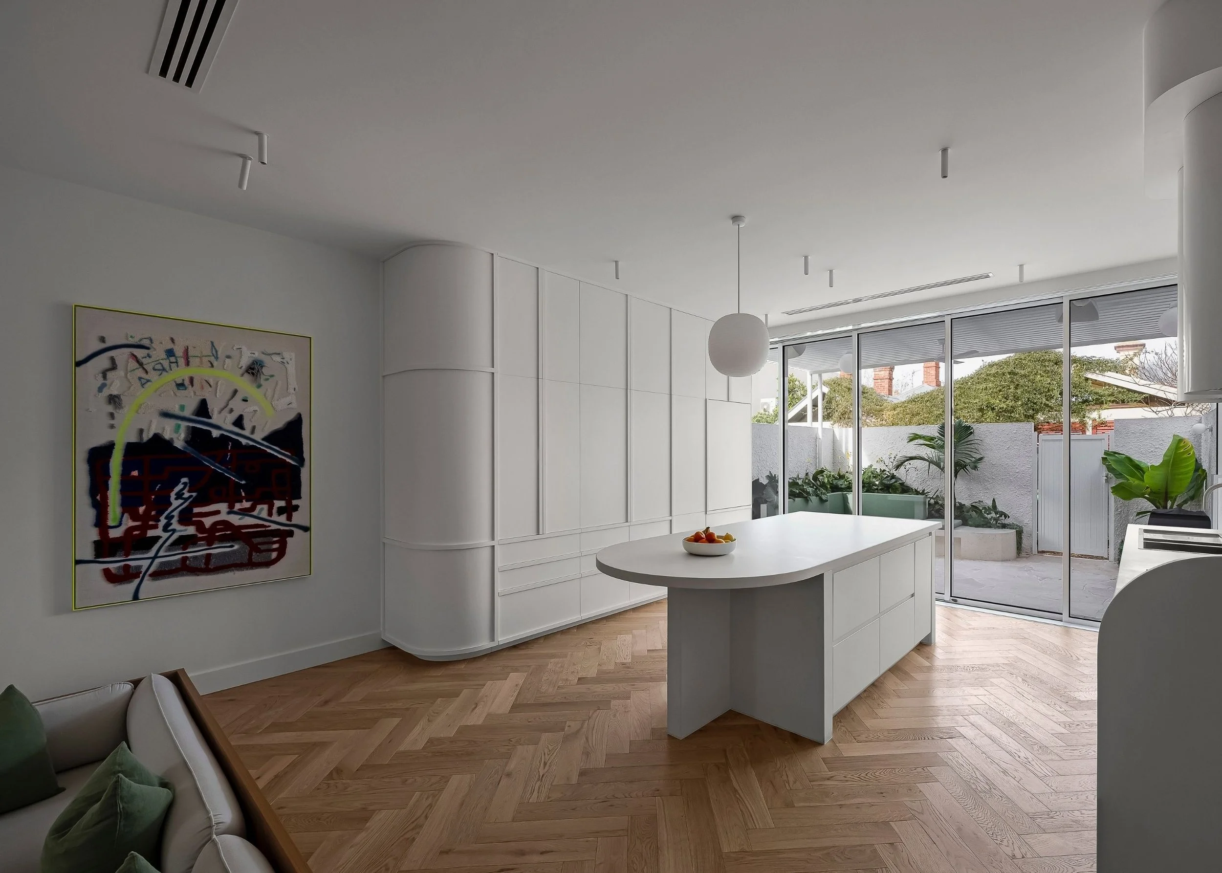 Modern living room with white cabinetry, a white oval table with a fruit bowl, green plants outside through sliding glass doors, and abstract artwork on the wall.