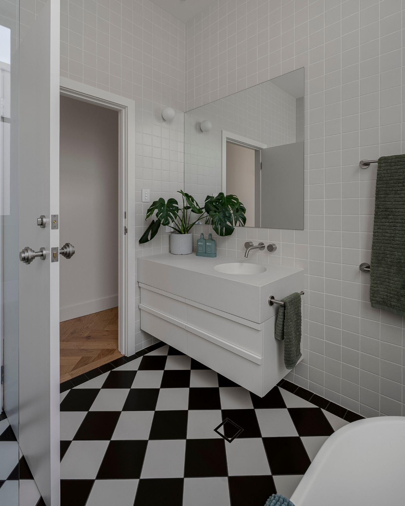 Modern bathroom with white tiled walls, black and white checkered floor, white vanity with a round sink, large mirror, potted plant, and gray towels.