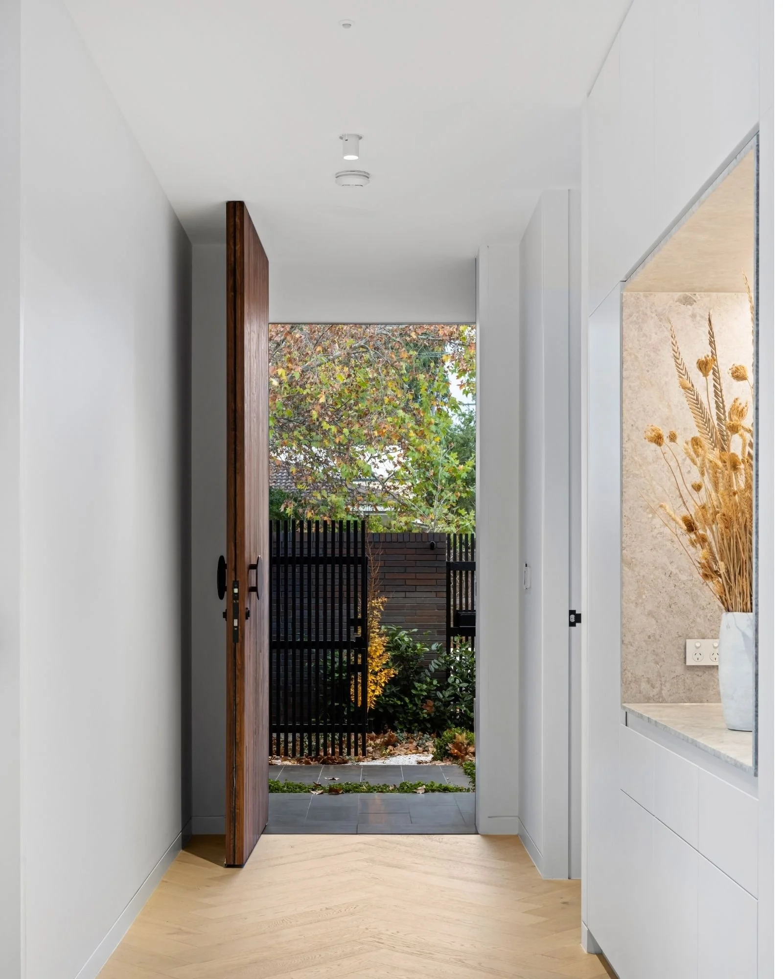 View through a modern home entryway showing a wooden door, white walls, and a patio with plants and trees outside.