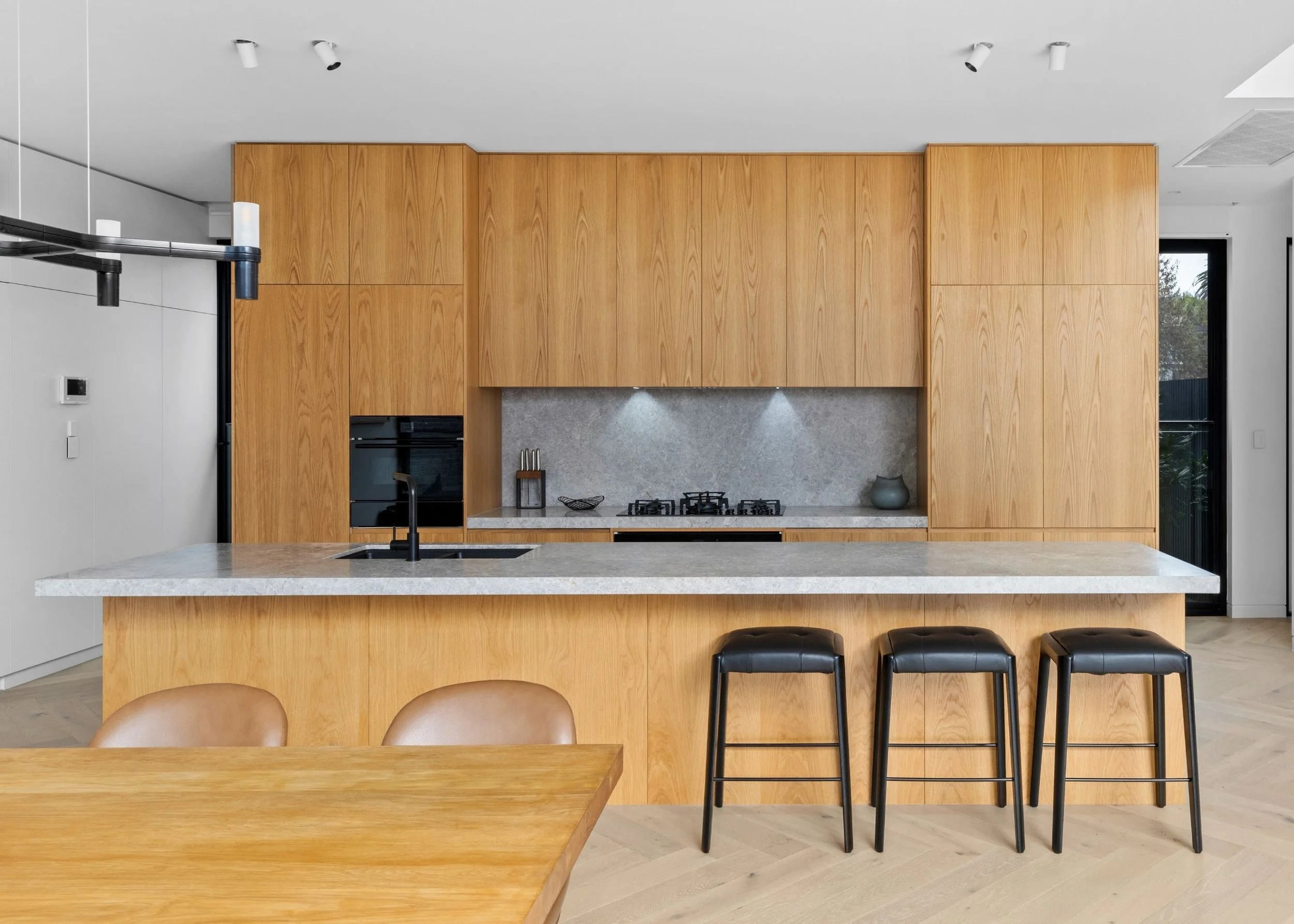 Modern kitchen with wood cabinets, a gray backsplash, a stone countertop island with stools, and a large window showing outdoor trees.