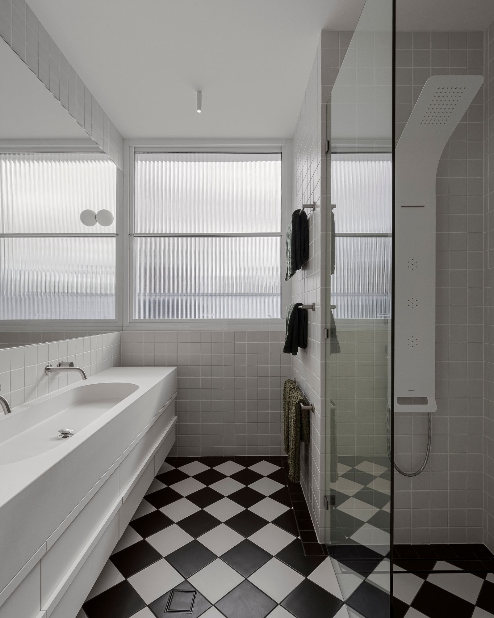 A modern bathroom with black and white checkered floor tiles, white wall tiles, a bathtub with a silver faucet, large windows with frosted glass, a towel rack with black towels, and a walk-in shower with a rainfall showerhead and a glass door.