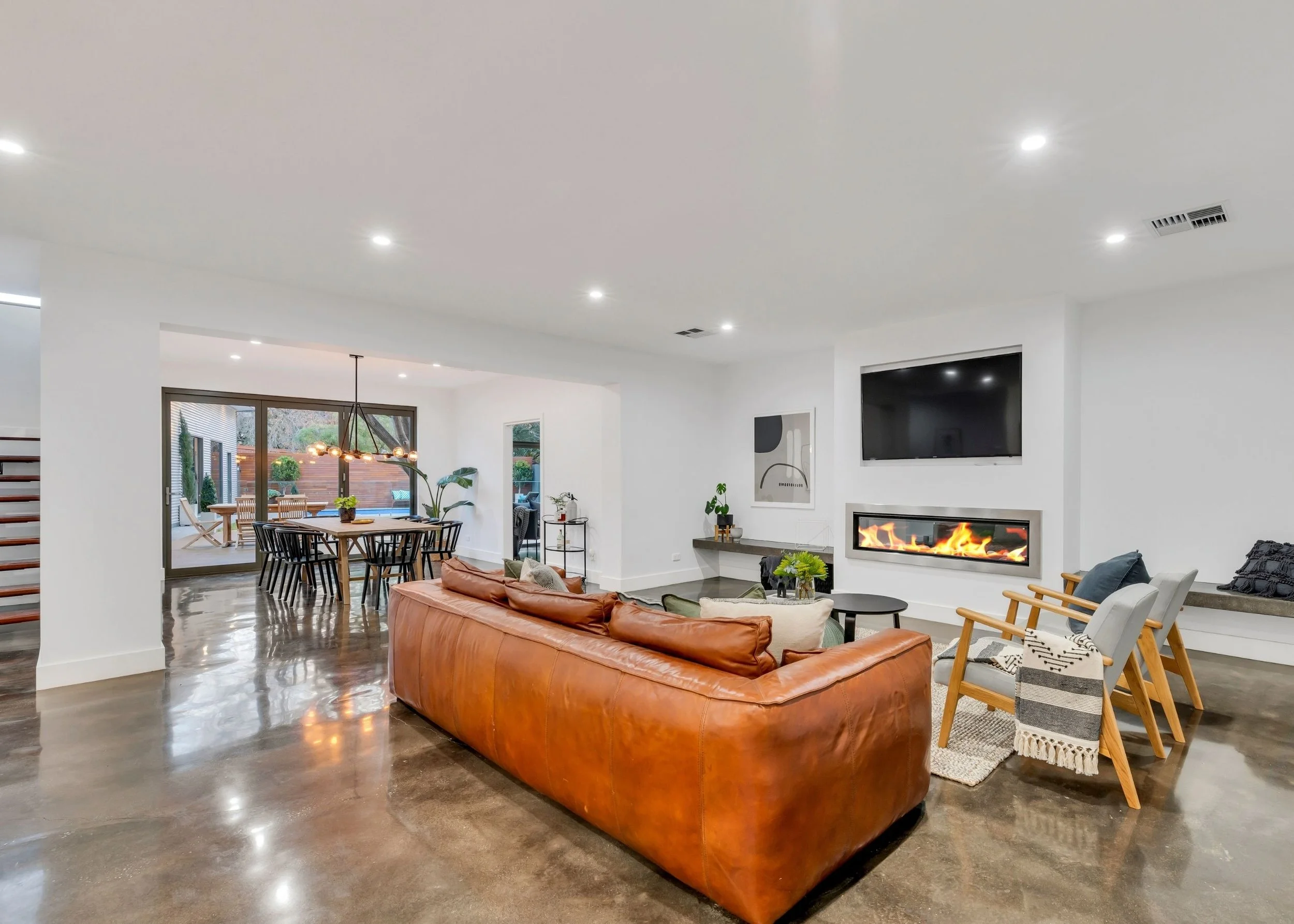 Modern living room with a brown leather sofa, a white armchair, a black TV mounted on white wall, and a fireplace with flames. Dining area with a wooden table, black chairs, and a chandelier, opening to an outdoor patio with trees.