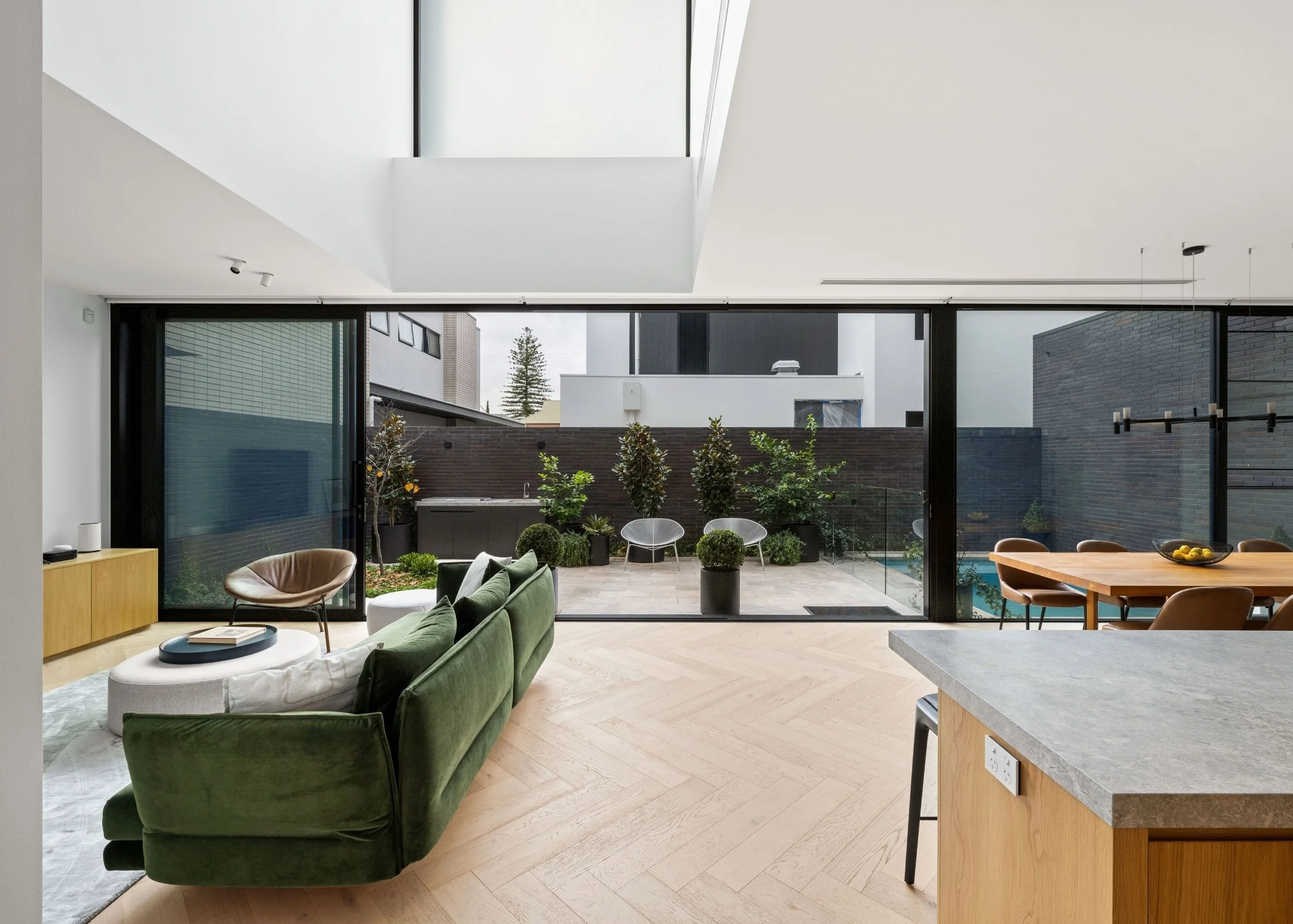 Modern living room with large glass doors opening to outdoor patio with plants and seating, contemporary furniture, light wood flooring, and a kitchen counter in the foreground.