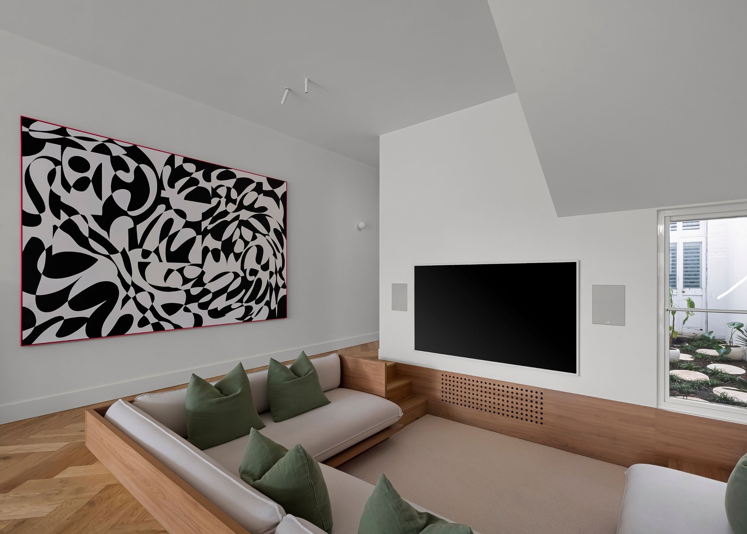 Modern living room with a white couch, green pillows, a black and white abstract painting on the wall, a fireplace, and a large window with a view of a garden.