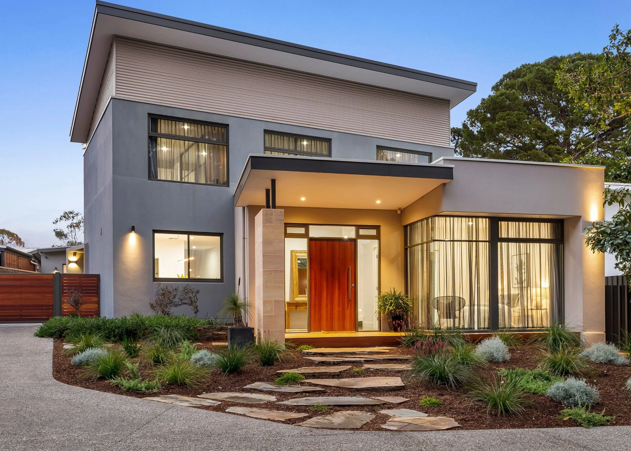 Modern two-story house with large windows, a front porch, and surrounding landscaped garden with pathway and trees in the background at dusk.