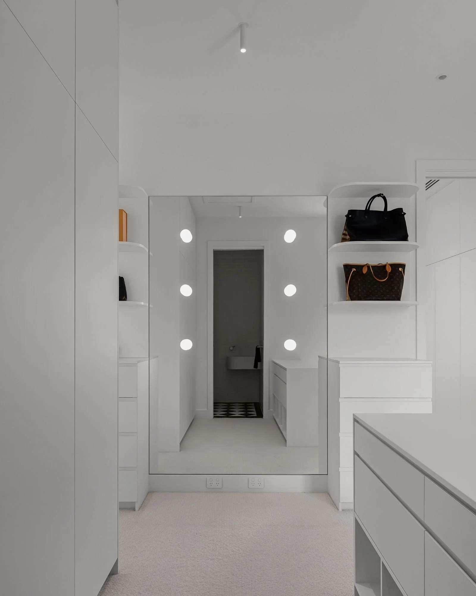 A modern, minimalist walk-in closet with white cabinetry, shelves, and drawers, featuring handbags on the right shelves, a mirror on the back wall, and ceiling lights.